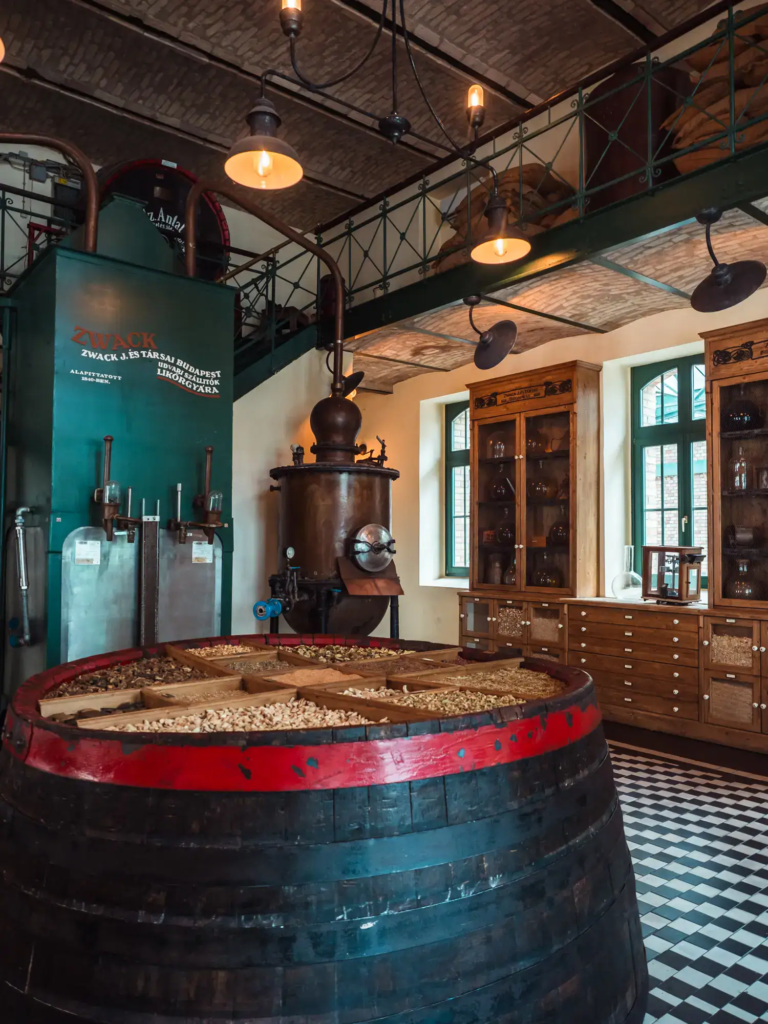 Inside the showroom with white and black checkered floors, old distillery equipment in the background, large black and red barrel with a tray of grains and spices on top, during a Zwack Unicum Factory Tour.