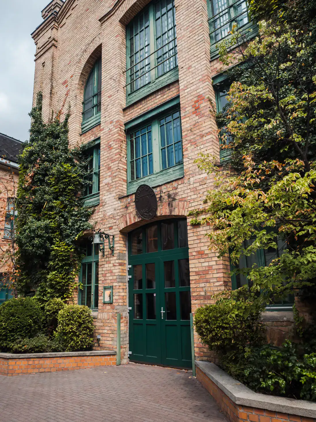 Side of large red brick building with green doors and windows, greenery growing on both sides, during Zwack Unicum Factory Premium Tour in Budapest.