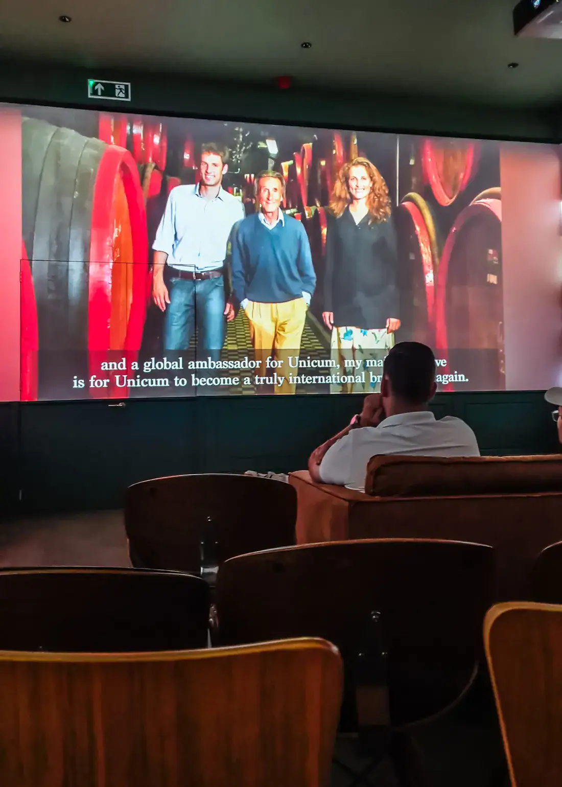 Inside small movie room with wooden chairs and the Zwack family on the screen in front, during a Zwack Unicum Factory and Museum tour in Budapest.