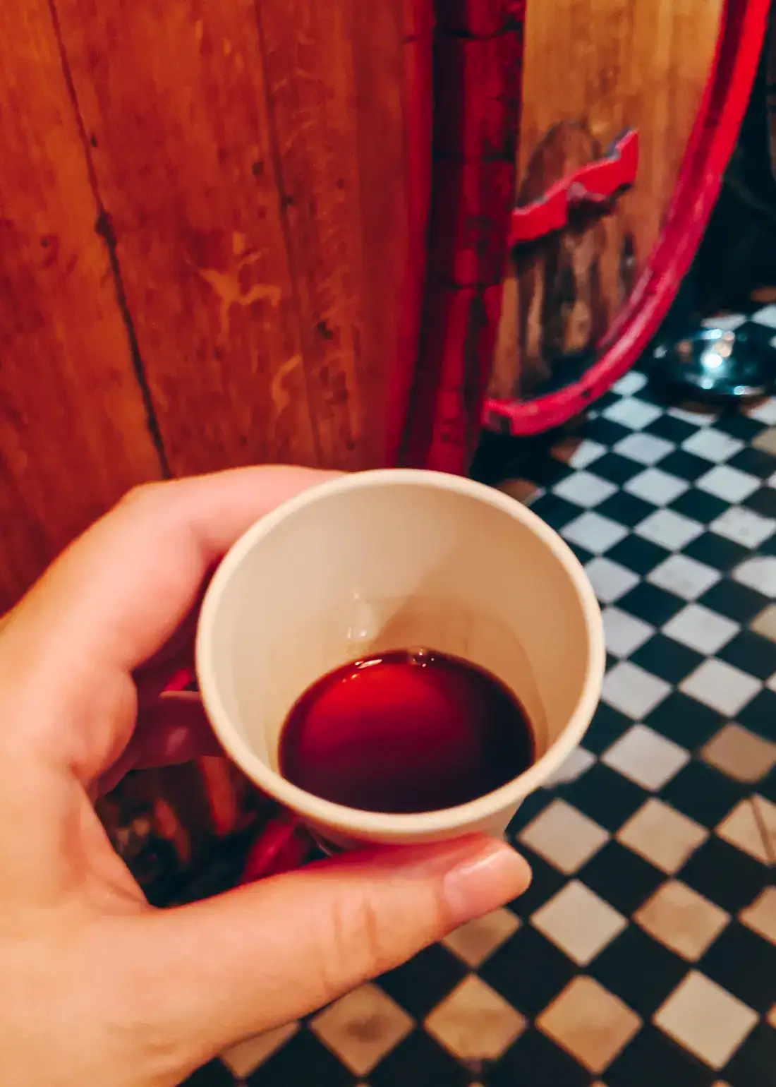 Hand holding a small, white cup with Unicum, wooden barrel and checkered floor in the background during a tour of the Zwack Unicum Museum and Factory in Budapest.