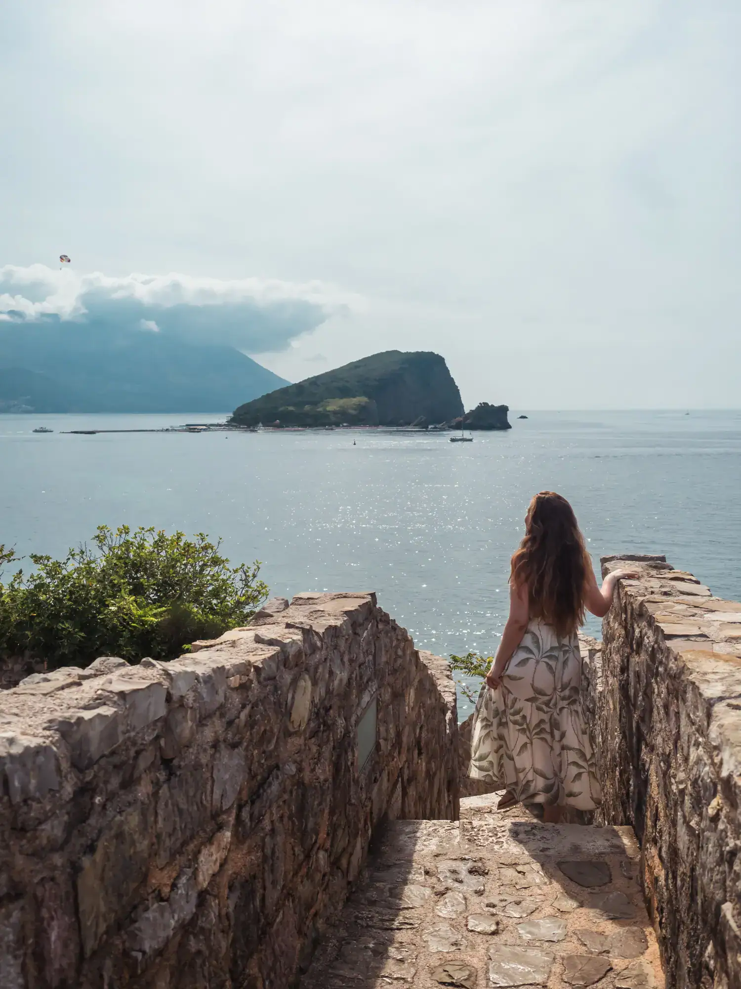 Woman with long brown hair, wearing a white dress with a green pattern, staring in the old stone stairs of Budva Citadel looking out over the ocean with an island and clouds in the background on a sunny morning.