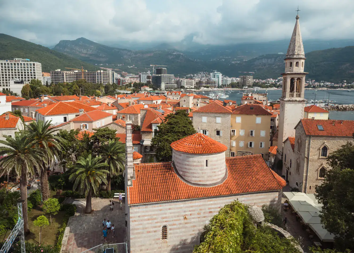 View from Budva Citadel across orange rooftops, a church spire and palm trees with Buda new town in the background. Where to stay, Budva or Sveti Stefan?