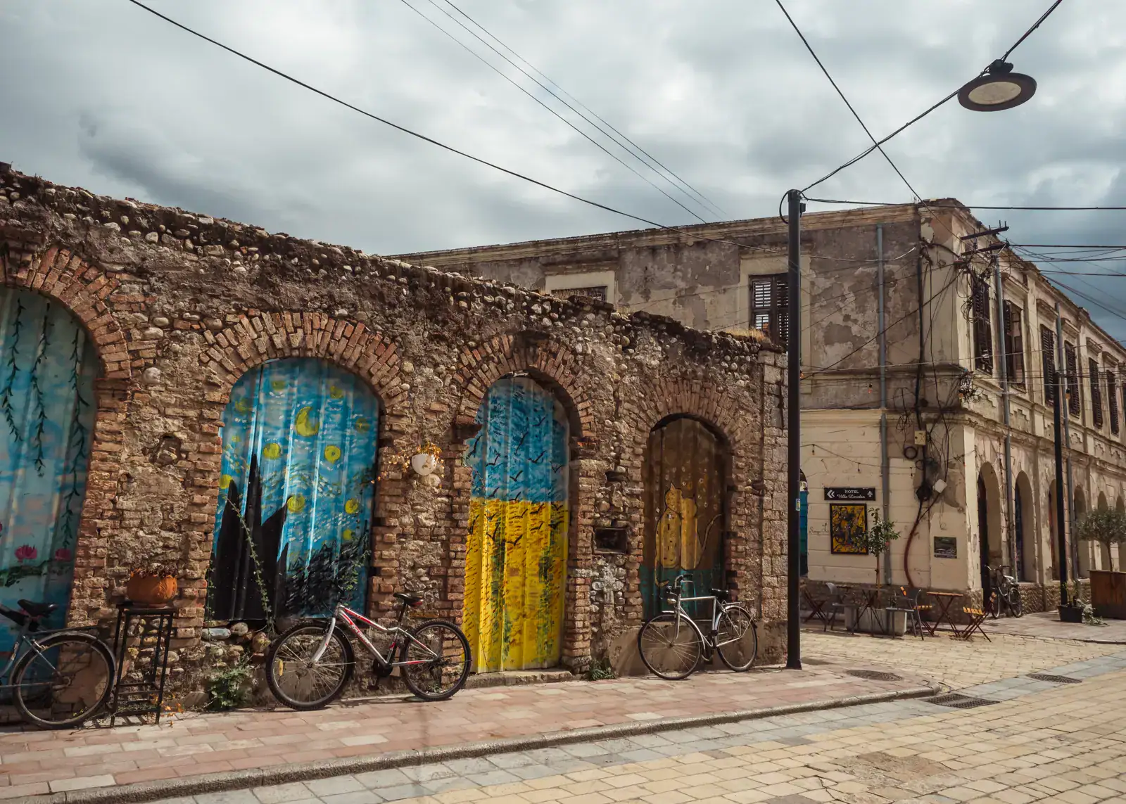Two bikes in front of a brick wall with arches with blue and yellow painted wood, old building in the background on a cloudy day in Shkoder.