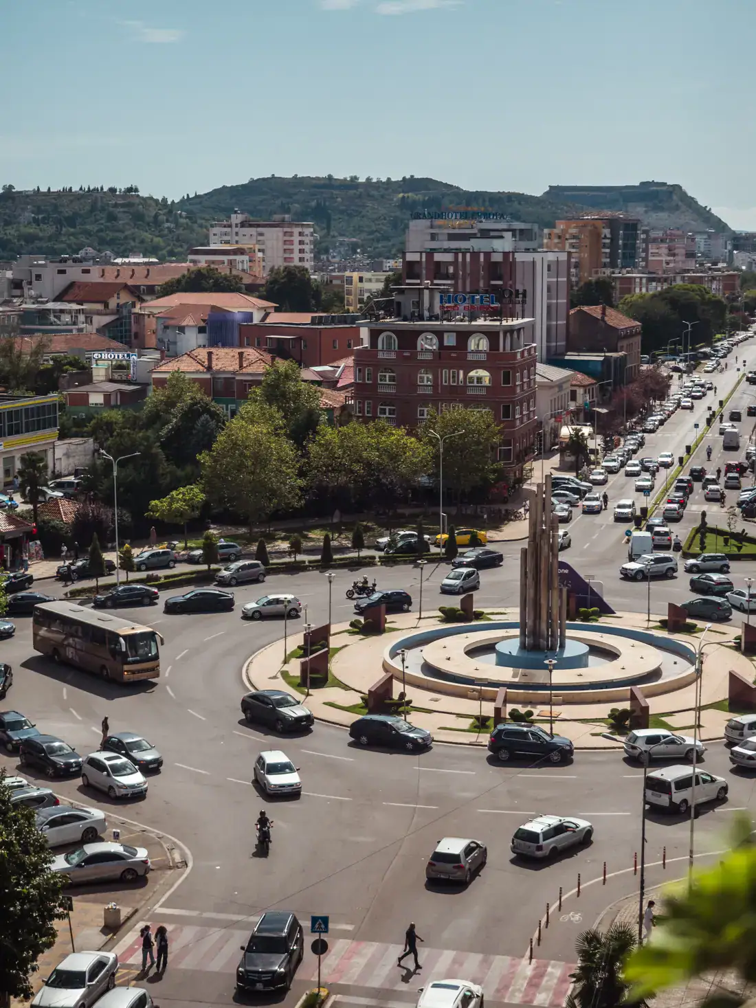 View from my balcony of the Main Street and roundabout in Shkoder with two lanes and parked cars on both sides, red, yellow and white apartment buildings in the background on a sunny morning.