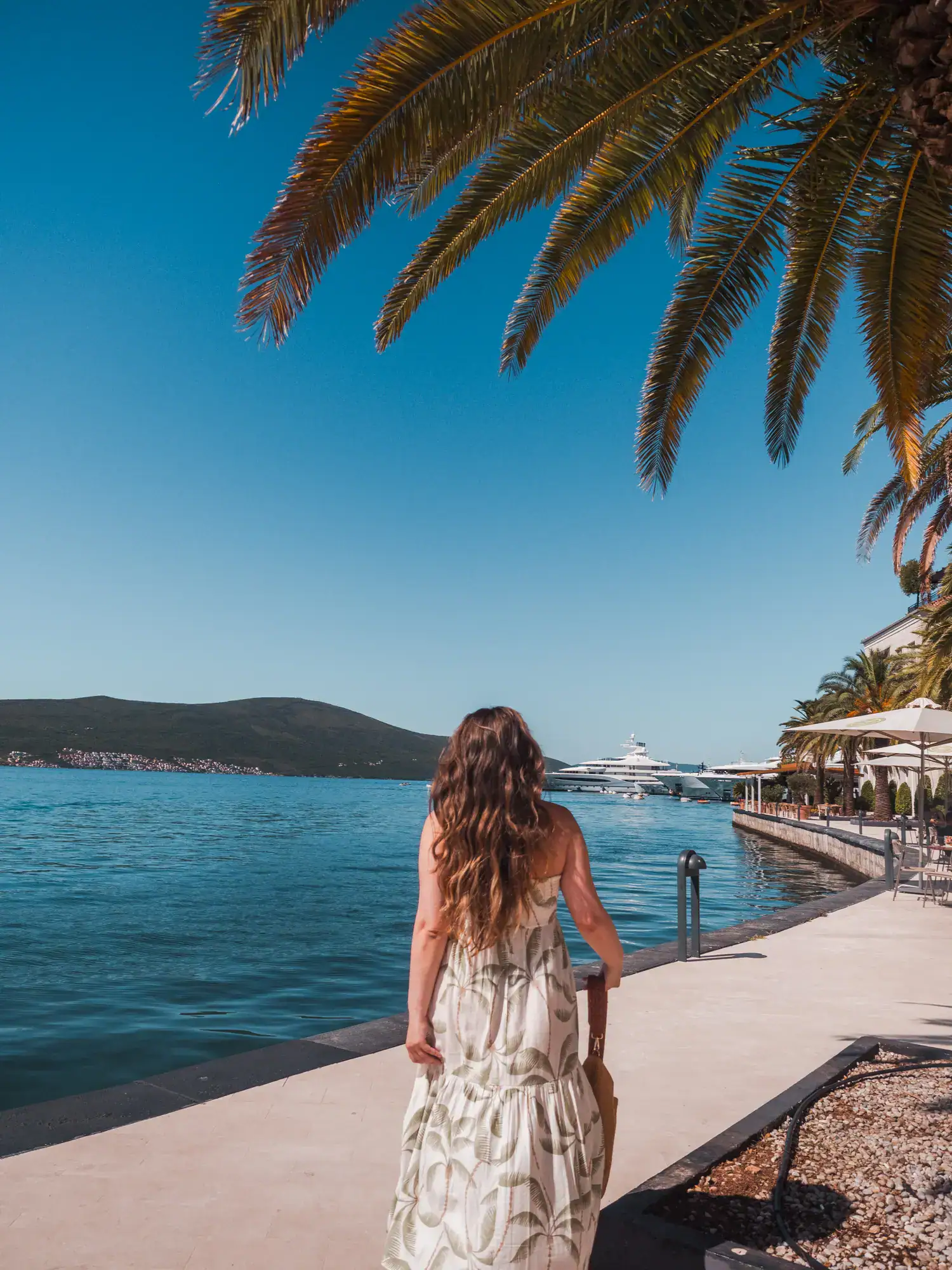 Woman with long, brown hair, wearing a white dress with green palm tree print, holding a brown bag, walking under a palm tree on the edge of Porto Montenegro, blue skies and water in Tivat.