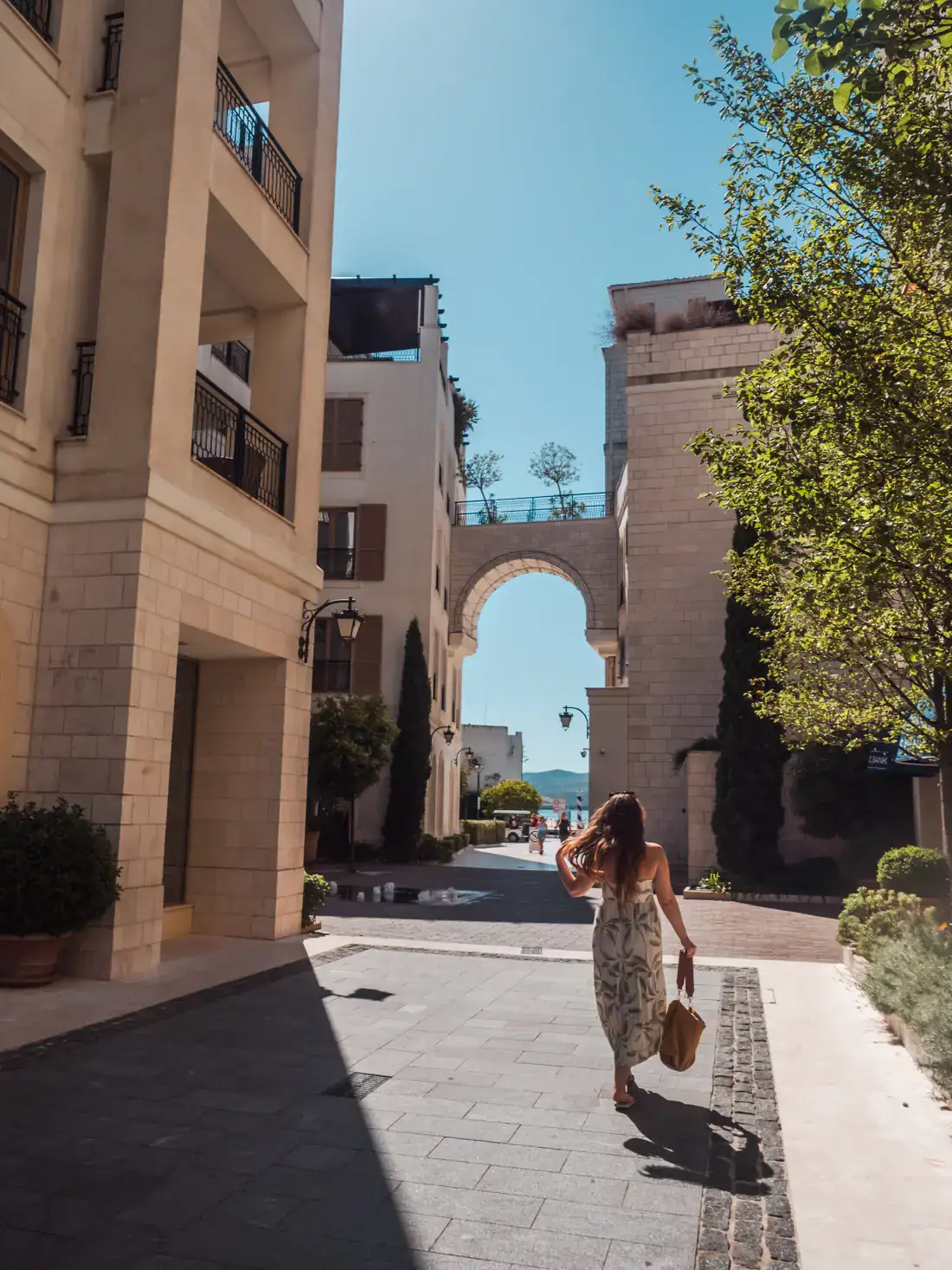 Woman wearing a white dresss, holding a bag and flicking her hair, walking towards an arch between to buildings, on a sunny day at Porto Montenegro in Tivat.