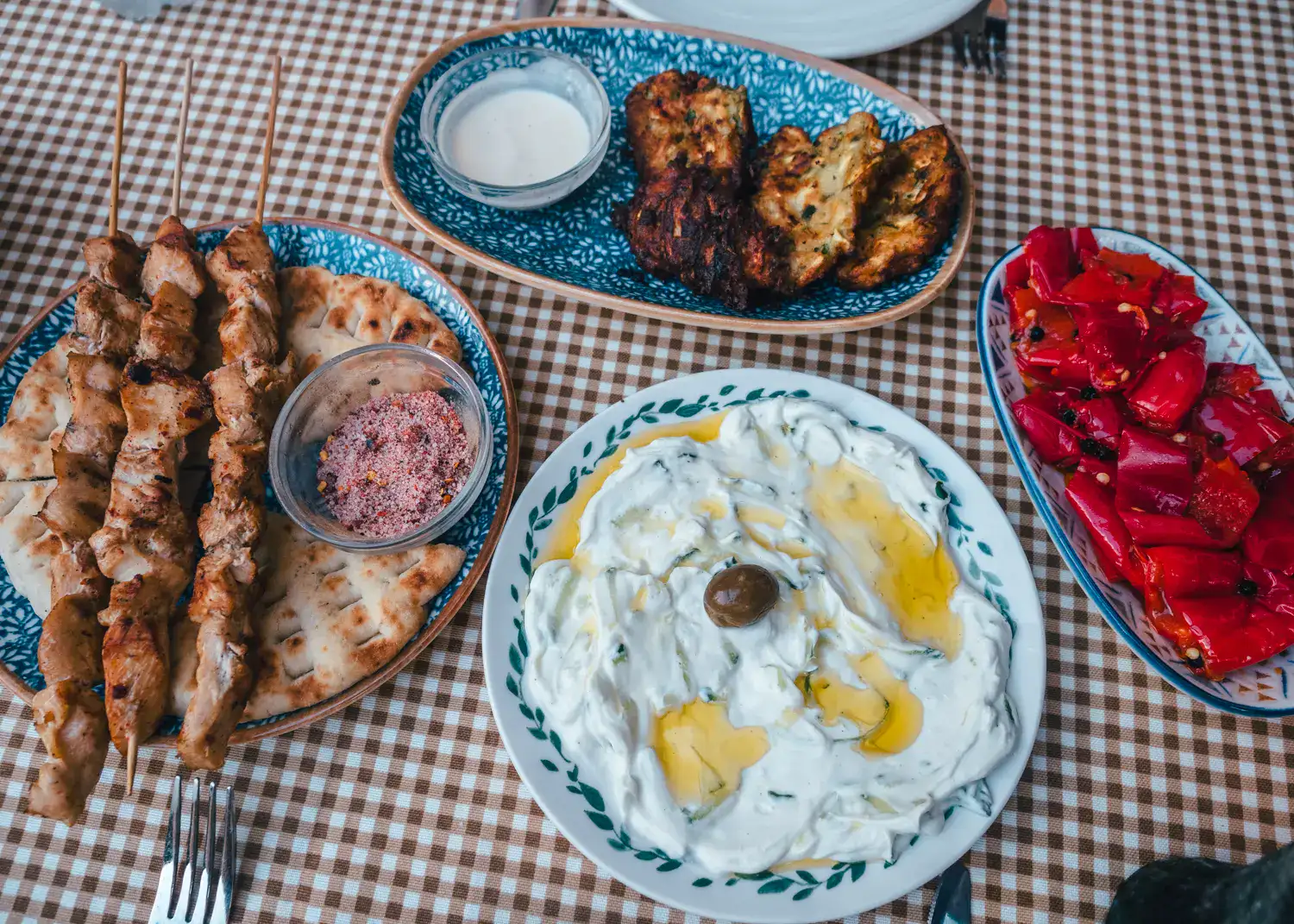 View from above of brown and white checkered table cloth, tzatziki with olive oil on a round plate, three chicken skewers, zucchini fritters, on a blue plate and grilled red peppers, at Mezzeria, one of the best restaurants in Shkod&euml;r.