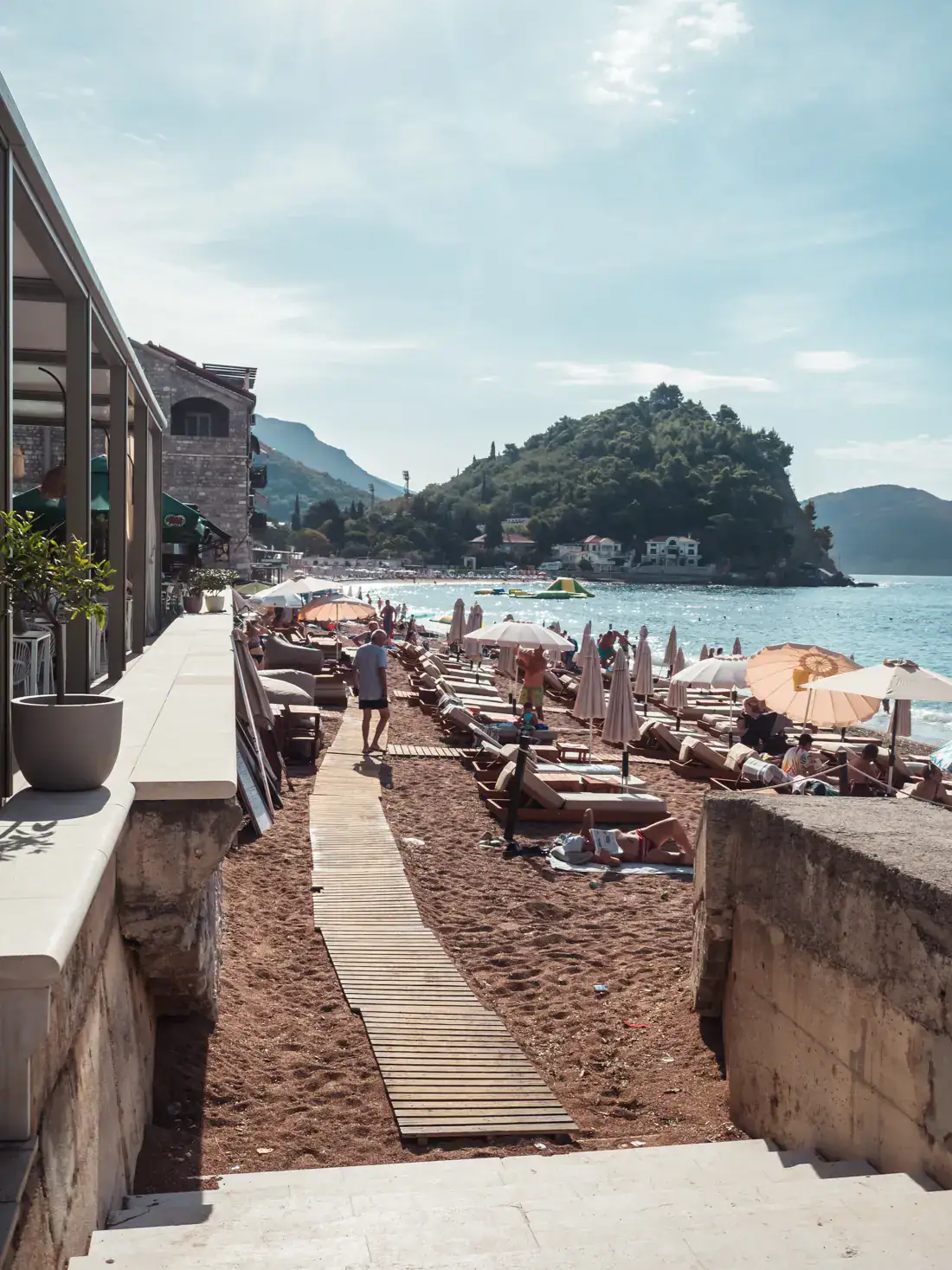 Wooden path on the sand on Petrovac Beach, lots of people on sunbeds, green island in the background, early morning in Petrovac.