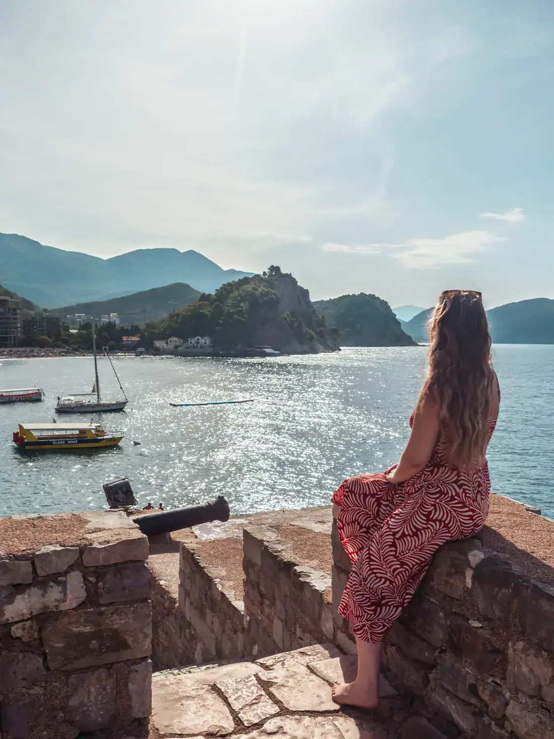 Woman in a red dress sitting on the edge of the old, stone Petrovac Castle, looking out over glistening ocean with islands and green hills in the background on a sunny morning in Petrovac.