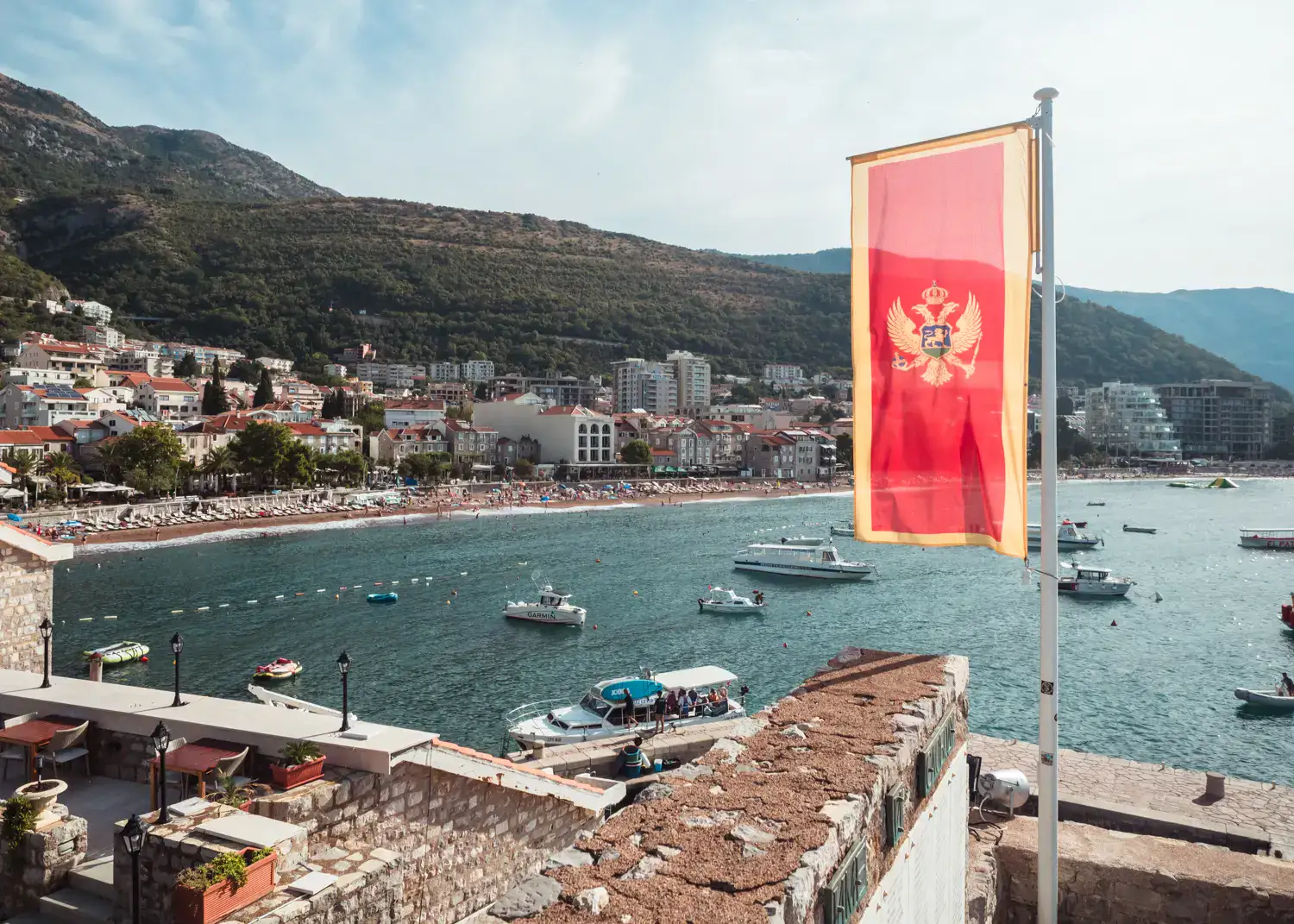 View from Petrovac Castle of the Montenegrin red and yellow flag in the wind, with the city beach and buildings in the background, many boats on the water. Where to stay, Budva, Sveti Stefan or Petrovac?