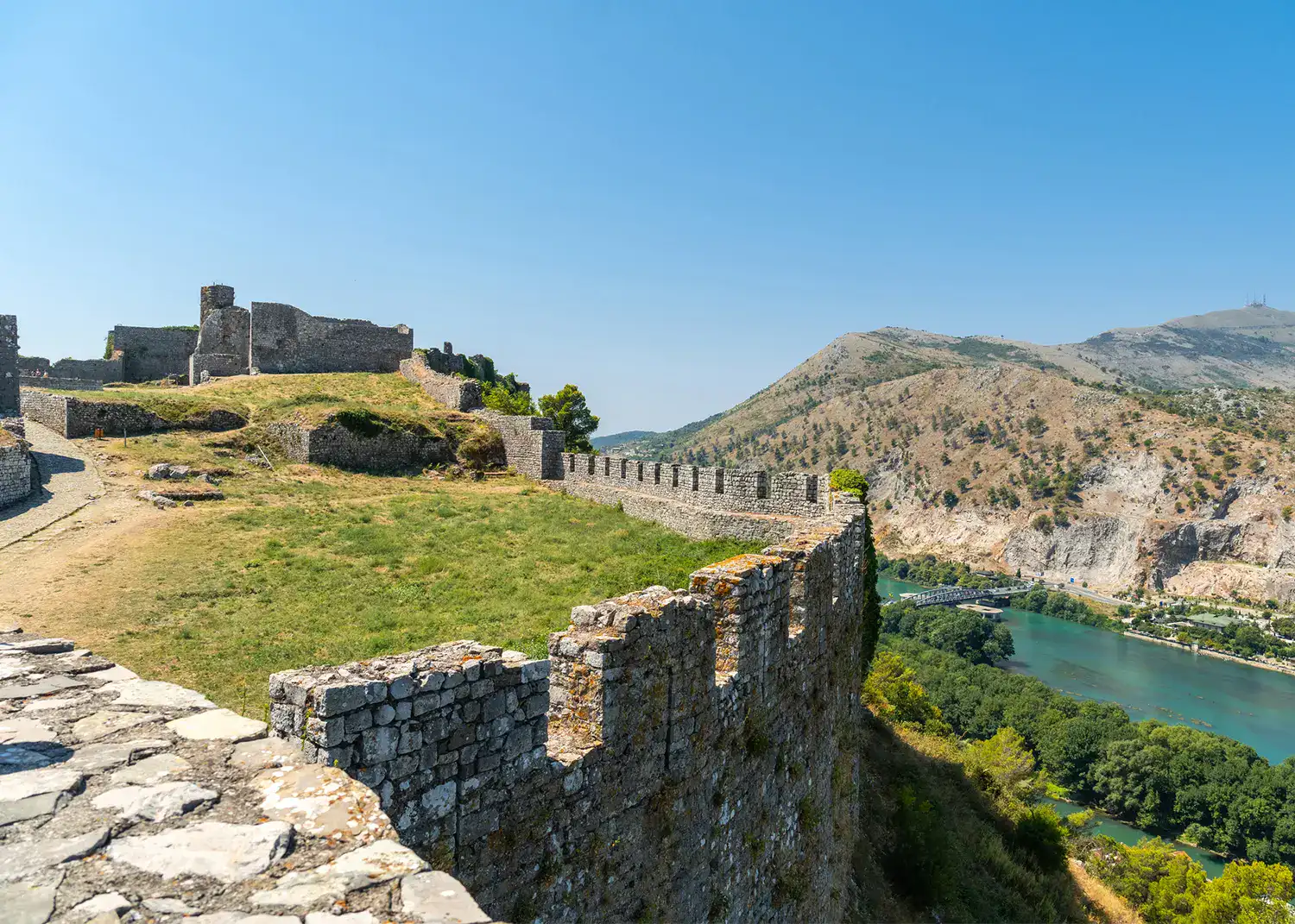 Rounded stone wall of Rozafa Castle with grass on top, turquoise river and greenery beneath, mountain in the background, on a sunny day in Shkoder, Albania.