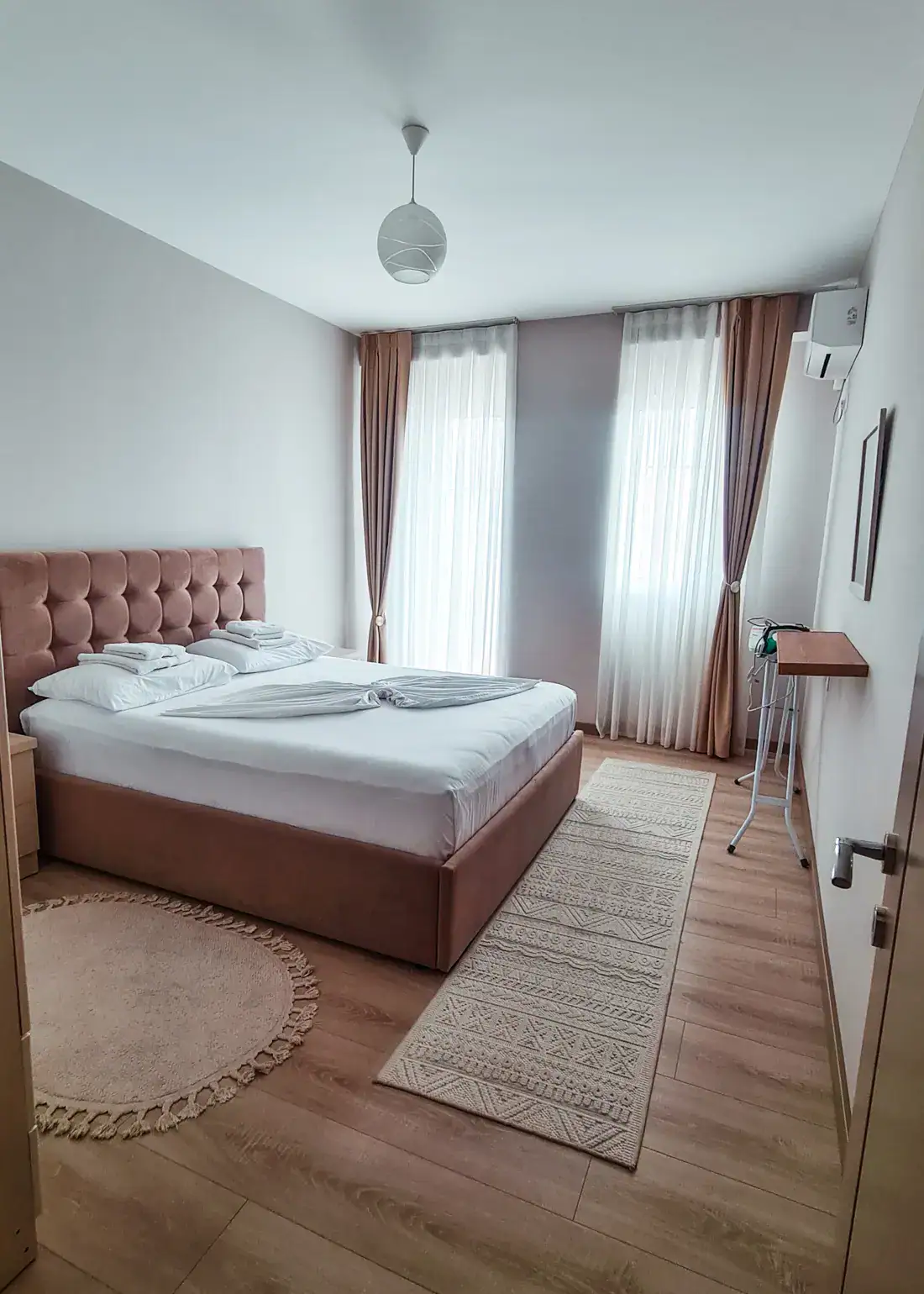Light brown bed with fabric headboard, white sheets and pillow cases, large windows covered with white curtains, ironing board on the right, best accommodation Shkoder.