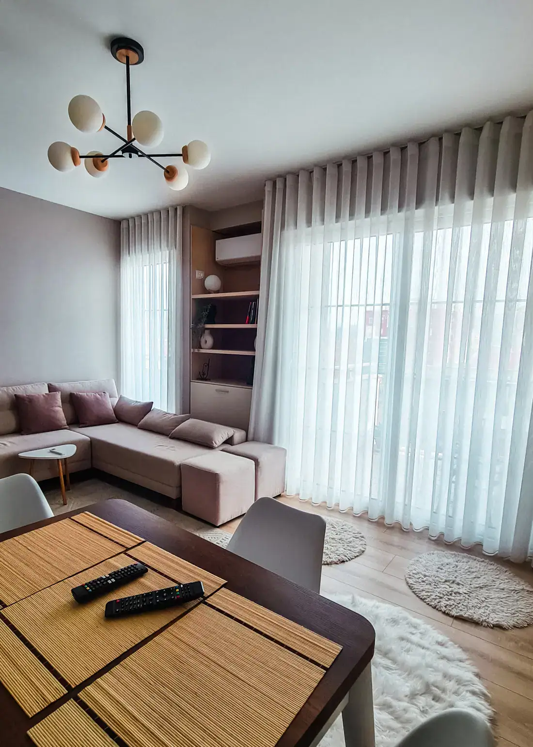 Living room with sliding door and window covered by white curtains, shelves in between, beige sofa and a small white table, dining table with straw place mats closest to the camera, affordable accommodation in Shkoder.