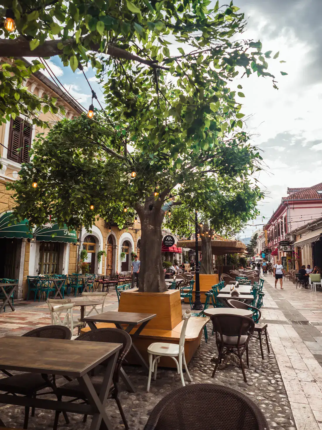 Tiled pedestrian street with buildings on both sides, trees in the middle with tables and chairs around it, is Shkoder worth visiting?