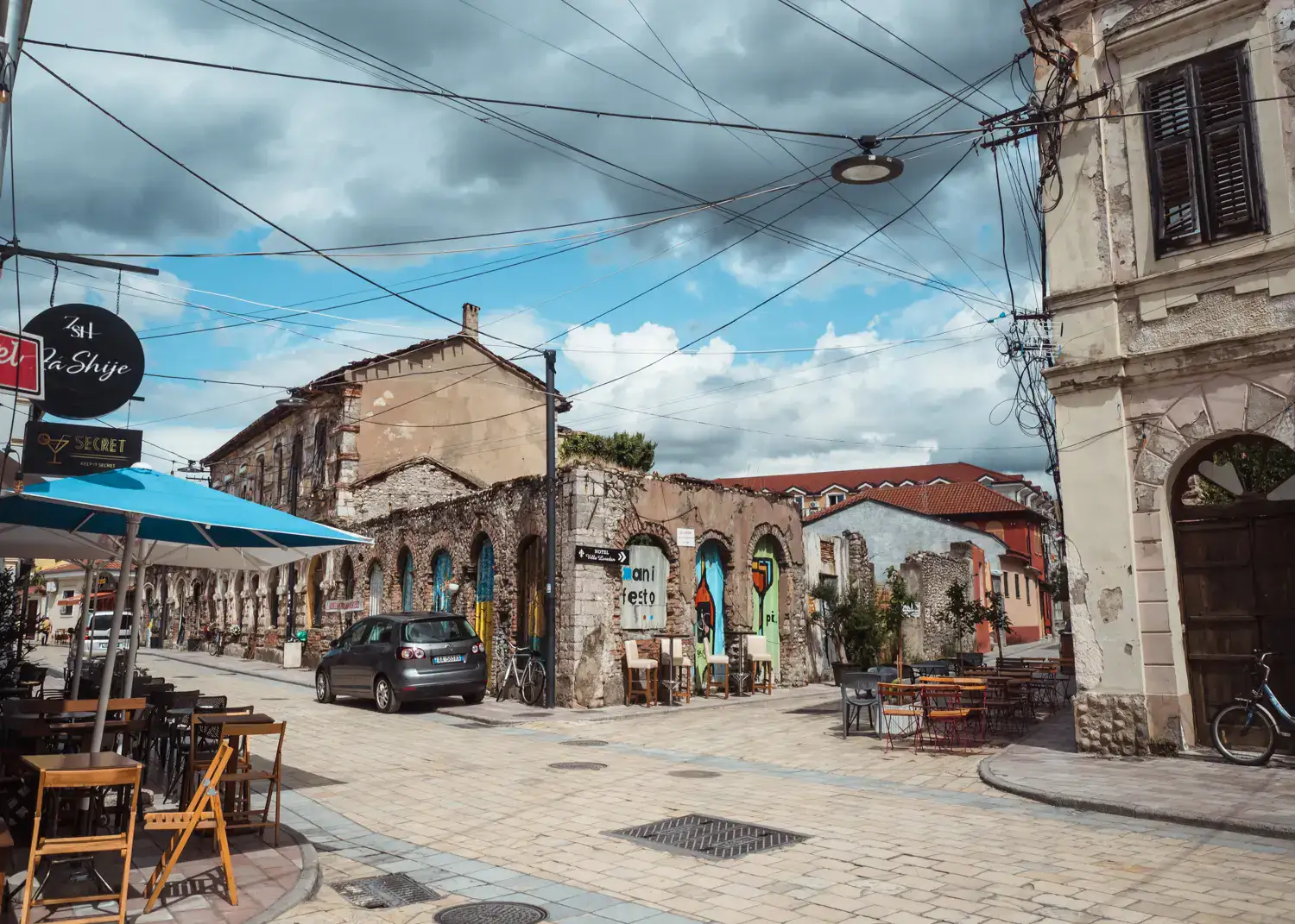 Tiles street, ruin building in the background with some street art, tables and chairs along the sidewalks, electrical wires crossing above on a cloudy day in Shkoder.