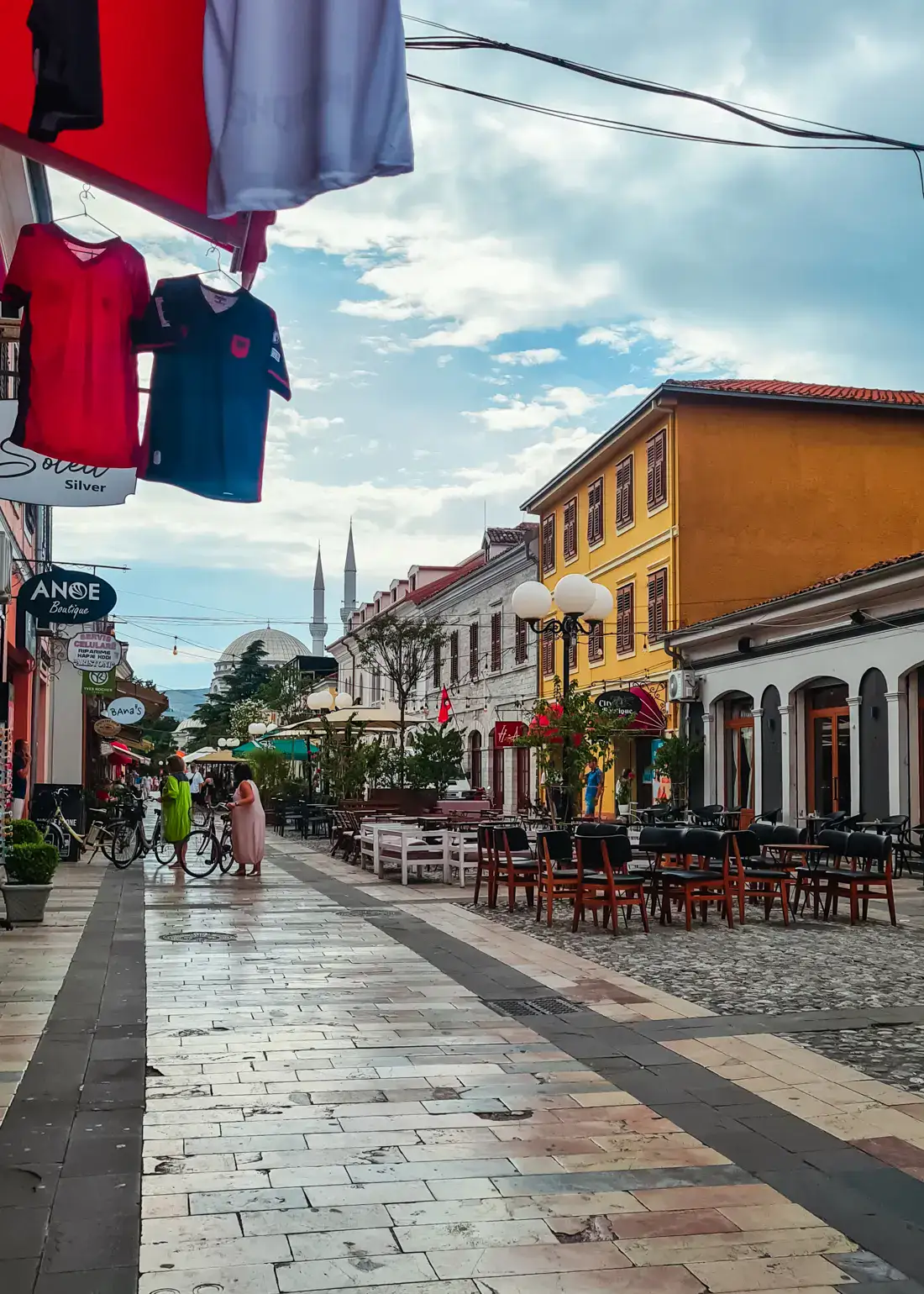 Tiled pedestrian street in Shkoder, shirts in a souvenir shop hanging on the left, yellow building and mosque spires in the background in the right, table in the middle.