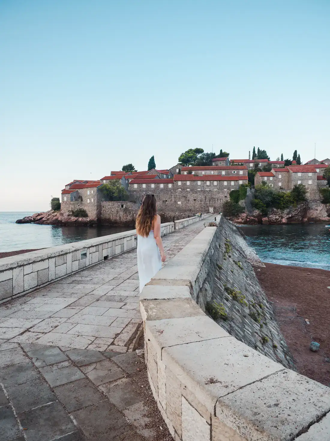 Woman with long brown hair, wearing a white dress, walking on the stone pathway out to Sveti Stefan island with orange roofed stone houses and trees, early on a clear morning in Montenegro.