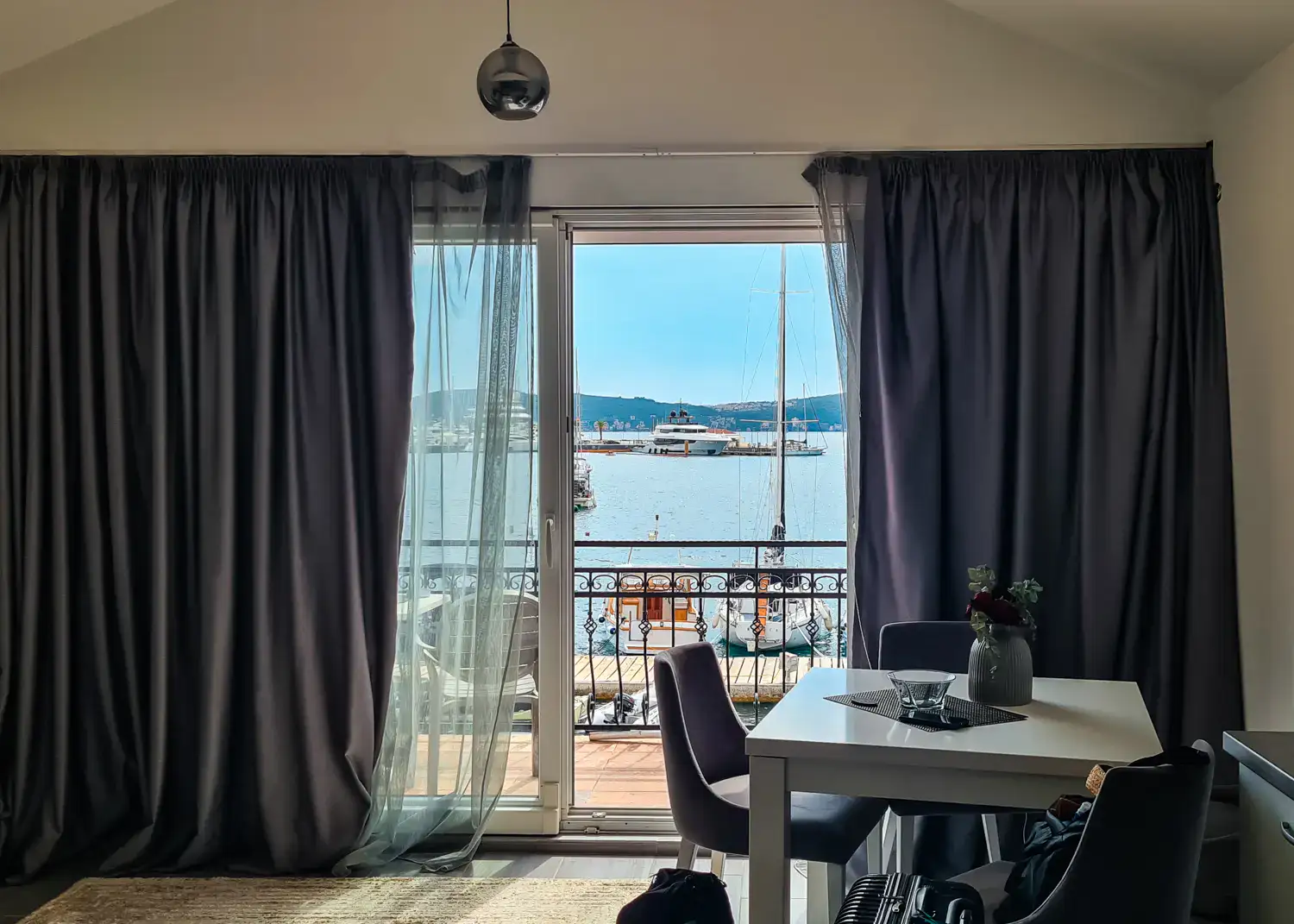 Looking through a living room with a white table and large sliding door, with purple curtains, onto the veranda overlooking Porto Montenegro in Tivat.