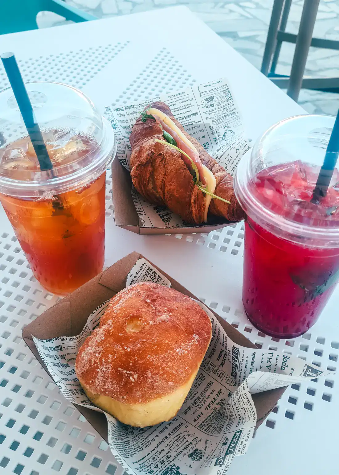 One iced tea, one red juice, one donut and one croissant with ham and cheese on a white table at one of the best bakeries in Tivat.