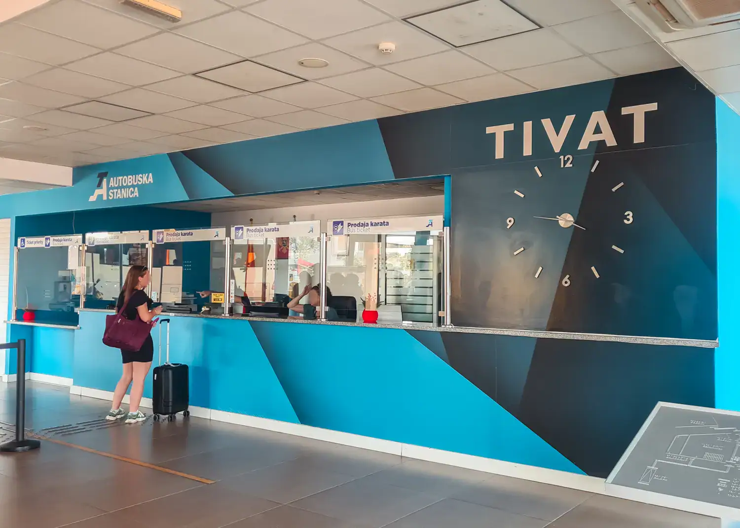 Tivat Bus Station, woman standing with a suitcase at the ticket counter on a wall painted in different shades of blue, with a large clock.