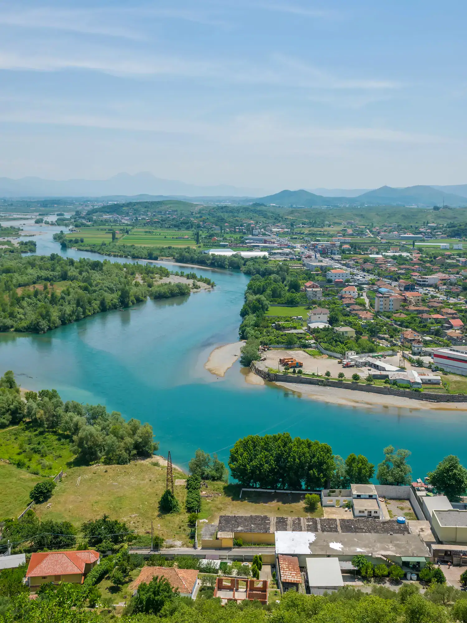 View of a turquoise river snaking between greenery and some buildings, seen from Rozafa Castle, early in the morning on a sunny day in Shkoder.