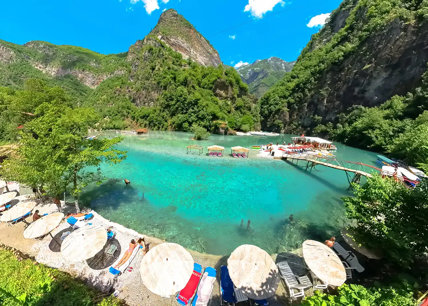 Wide angle view of the turquoise Shale river, mountains and greenery in the background, row of umbrellas in the foreground on a sunny day, visiting from Shkoder.