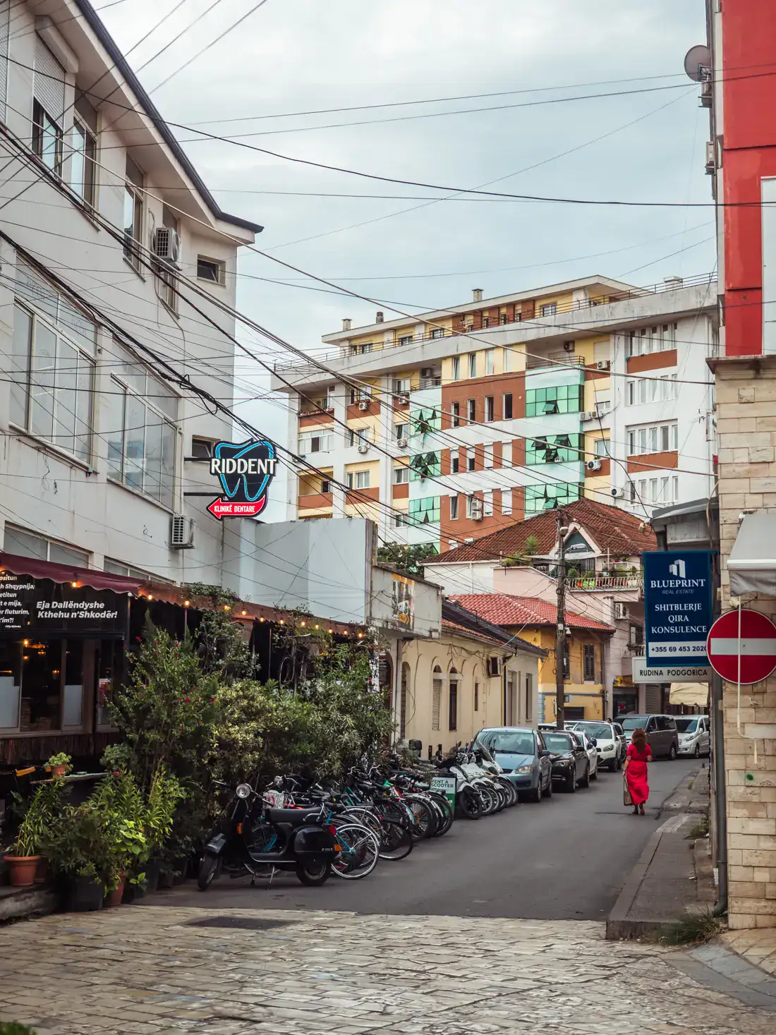 Woman in a red dress walking down a street surrounded by residential and commercial buildings in Shkod&euml;r Albania, with bikes and cars parked on the right and electric wires hanging above, on a cloudy day.