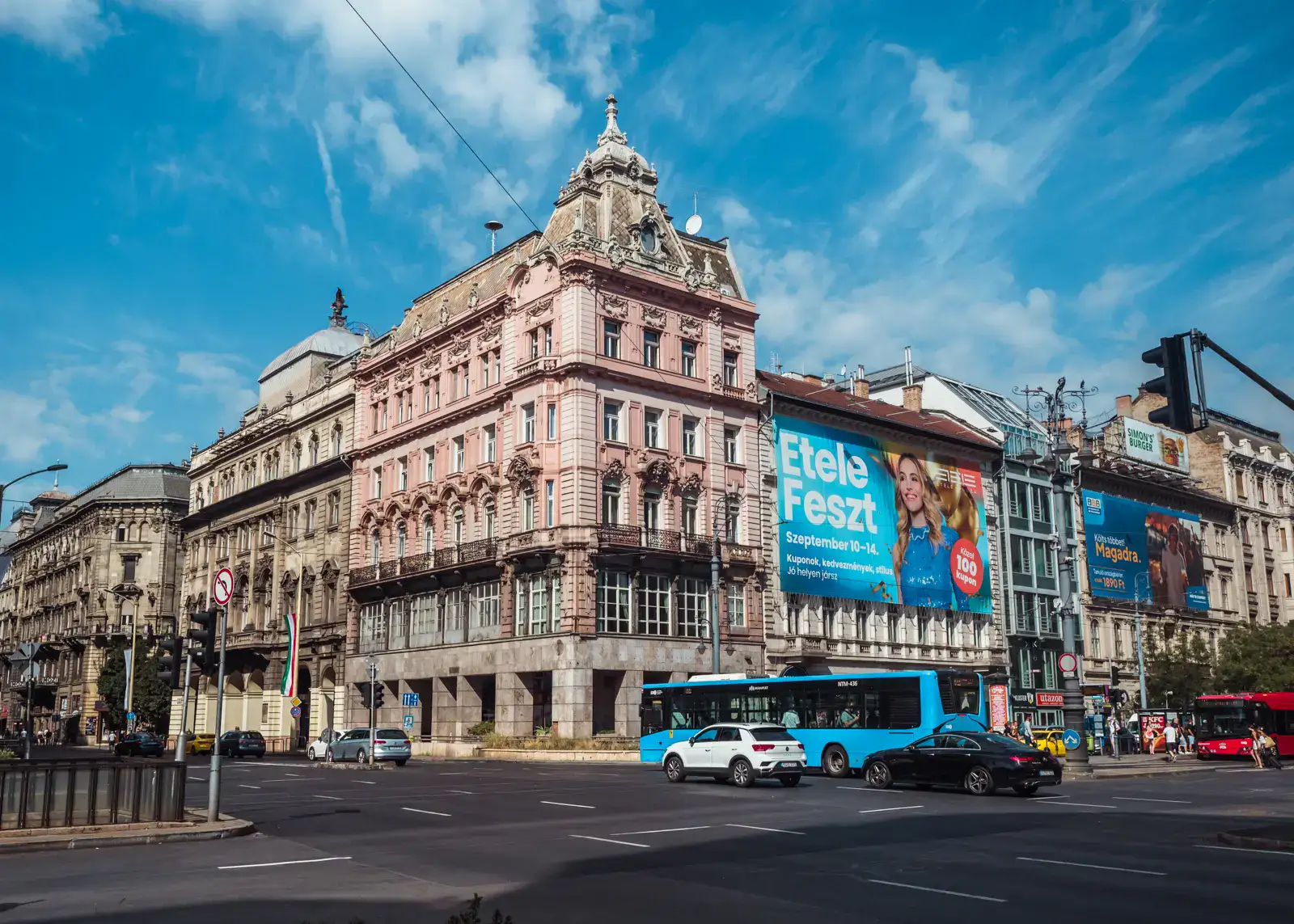 Large intersection in the center of Budapest, large, ornate, light pink apartment building in the background with a large blue ad on the right side, blue bus and a black and a white car driving across on a sunny day in Budapest.