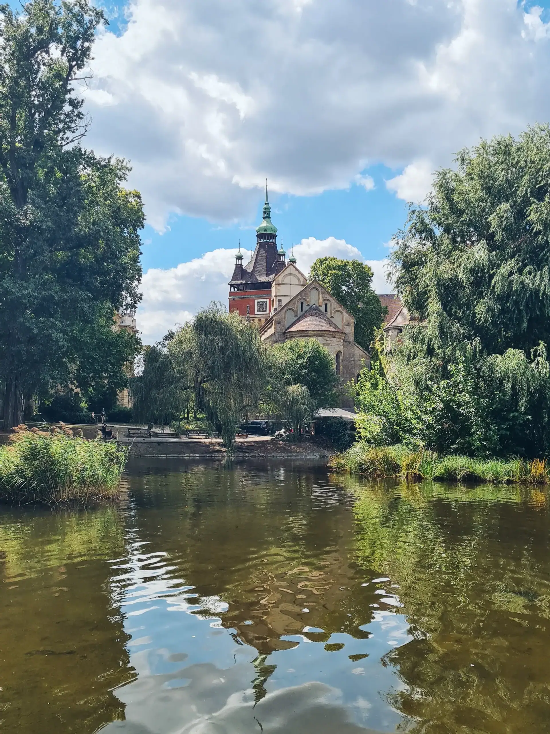 Looking over the water moat in the park around Vajdahunyad Castle, white church and red brick tower with a black roof and green dome seen through the trees, a must during your 3 days in Budapest.