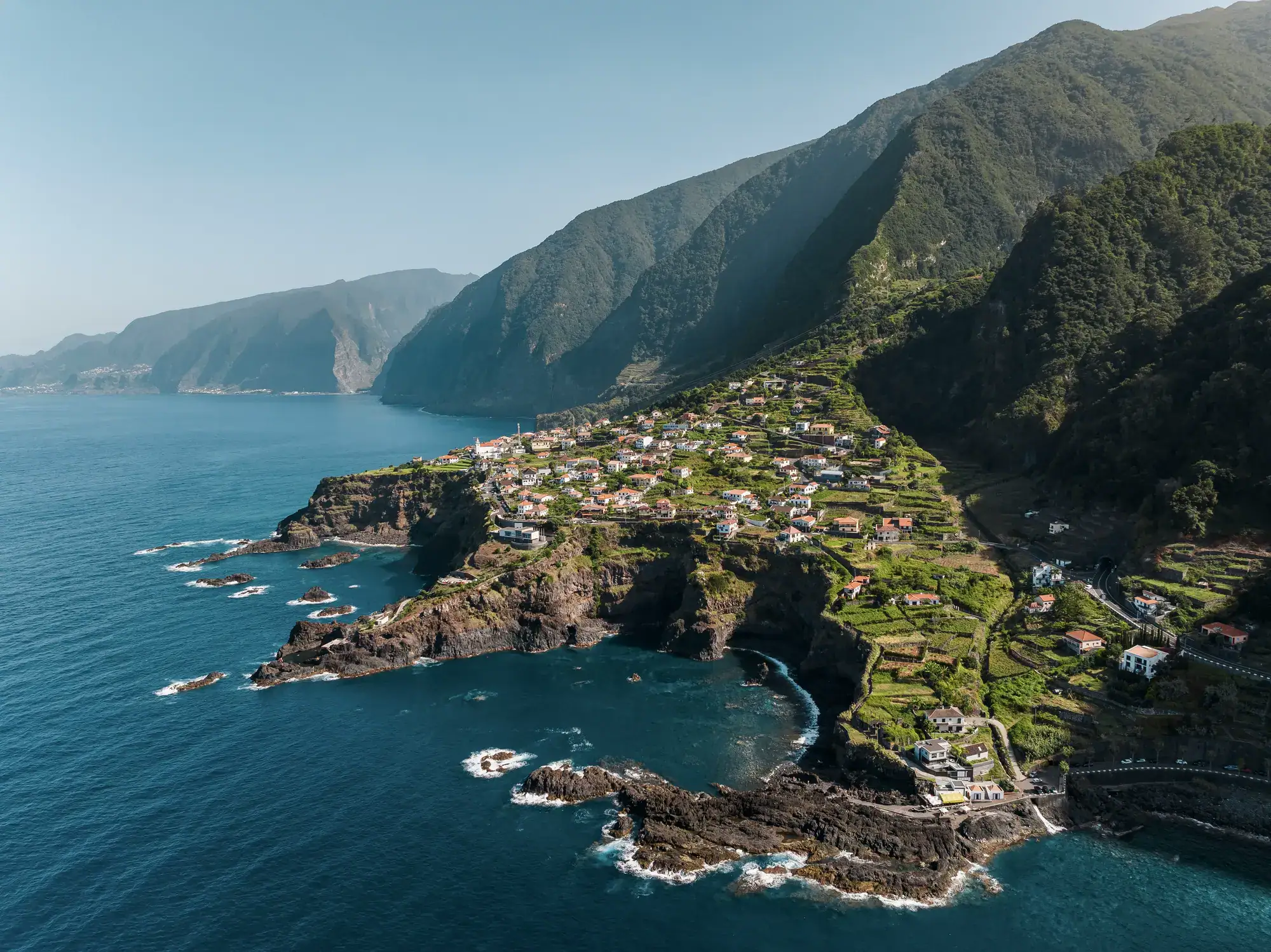 Seixal, a town set on a green, rugged cliff going into the dark blue water, mountains in the background, on a sunny day in Madeira, one of the warmest destinations to visit in Europe in December.