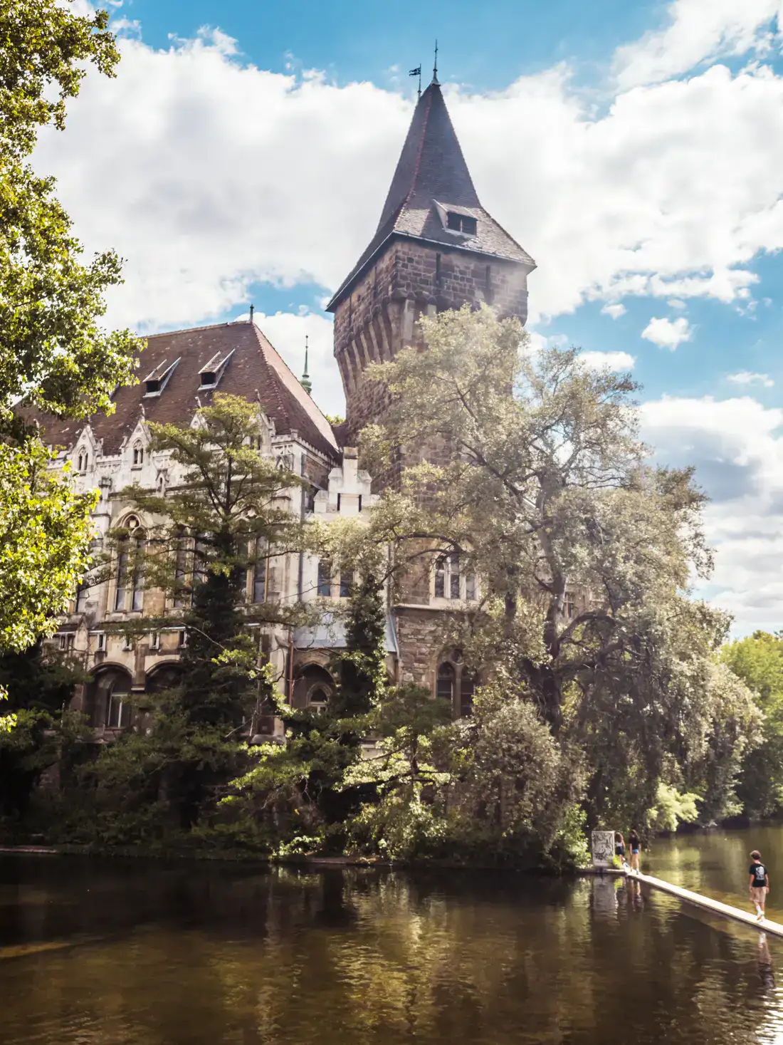 White and brown Vajdahunyad Castle, with a tower, behind green trees glowing in the sunshine, two people walking across the water moat on a thin path, on a lightly cloud day in Budapest.