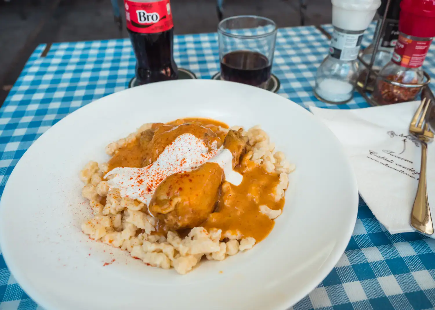 White plate with traditional Hungarian Chicken Paprikash, small dumplings with a chicken thigh and orange sauce, white and blue checkered table cloth, small glass coke bottle with "bro" on it. A must on any Budapest itinerary.