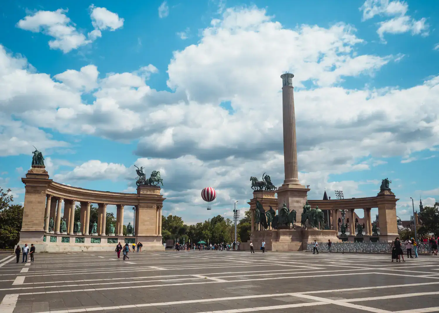 View of Heroes' Square with a tall pillar with one curved wall on each side on a tiled square with a red hot air balloon in the distance on a lightly cloudy day, 3 day Budapest itinerary. 