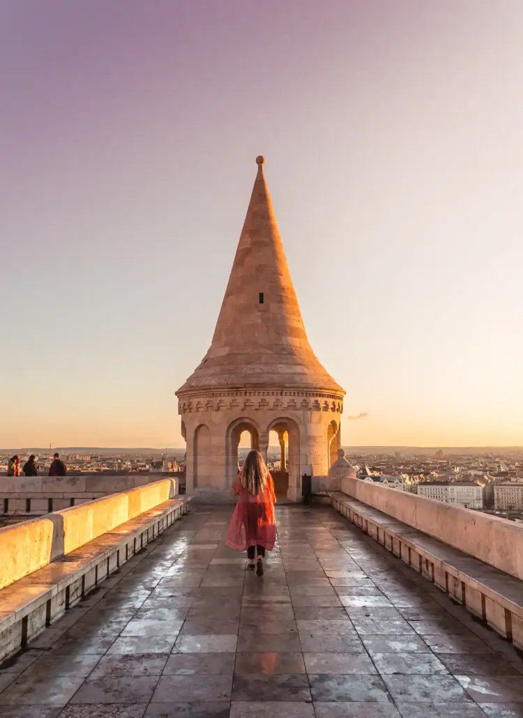 Woman with long hair, wearing black pants and a long, orange kimono, walking towards one of the sandstone turrets at Fisherman's Bastion during a light purple and yellow sunrise, a must on your Budapest itinerary.