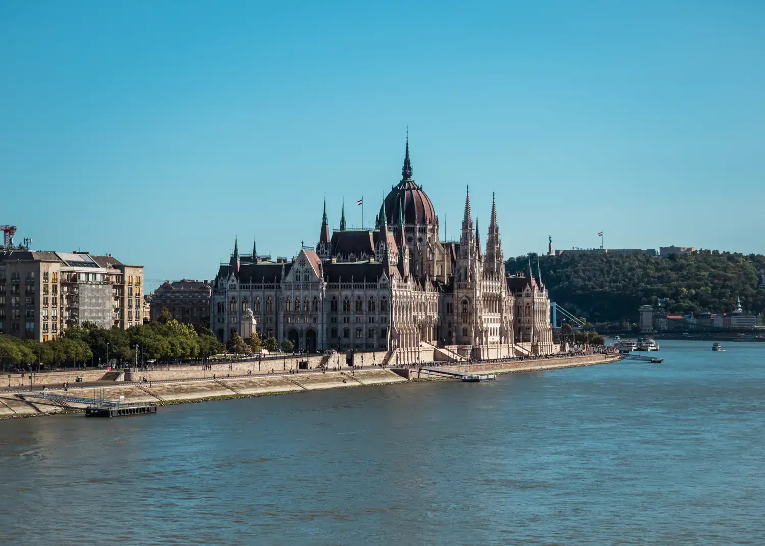 Looking from a distance, across the river, at the impressive, Gothic Hungarian Parliament with spires and a large done on the river bank, blue sky in the background. 3 day Budapest itinerary.
