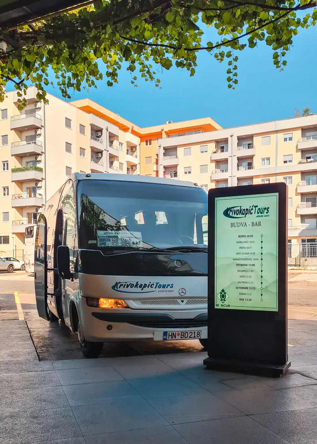 A furgon, small, white bus, by a digital sign at Budva bus station with a light yellow apartment block in the background, traveling from Budva to Tirana by bus.