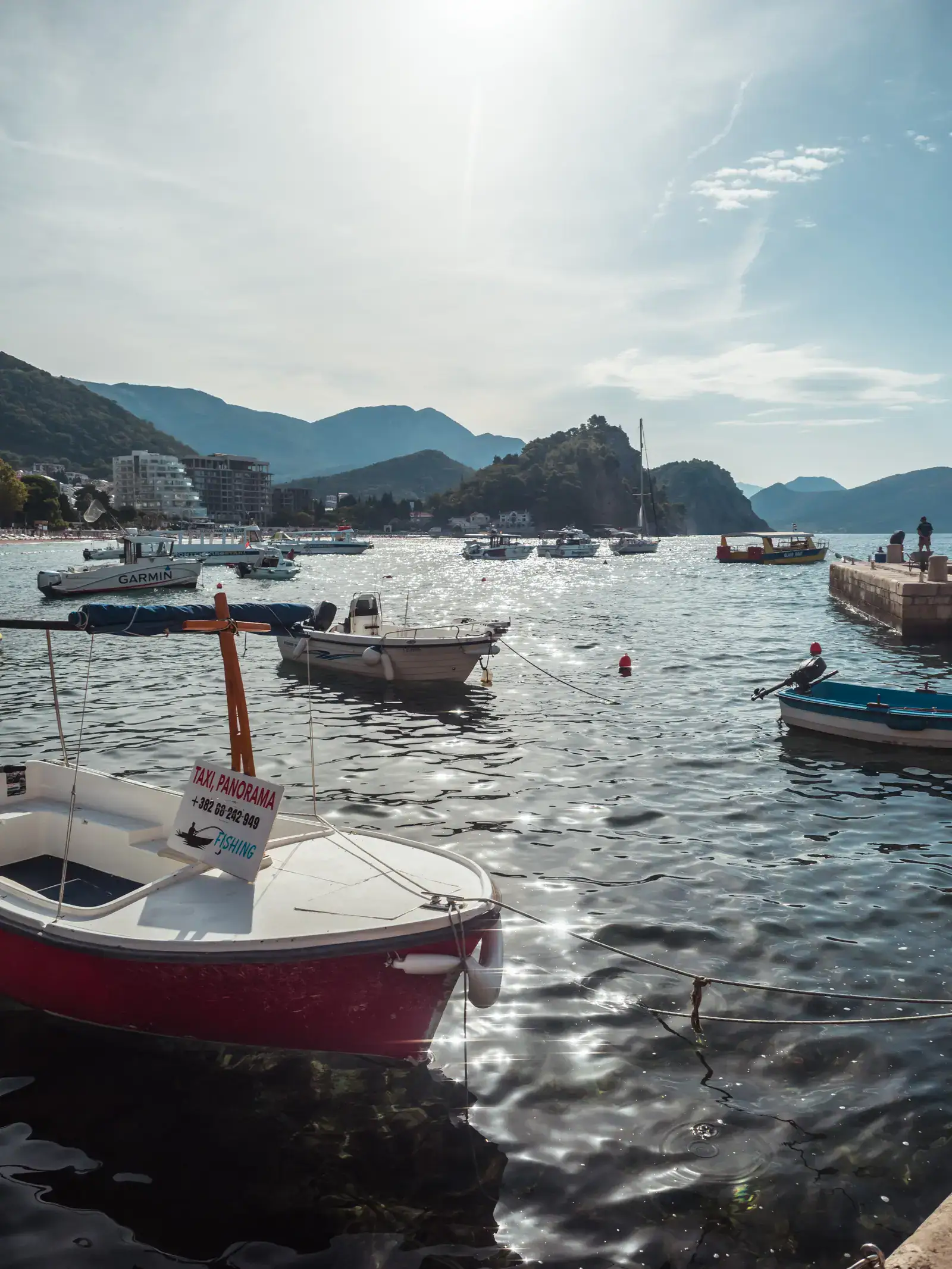 Several boats, one red in the foreground, on the glistening water in Petrovac Harbor with mountains in the background, on a stop between Budva and Tirana by private driver.