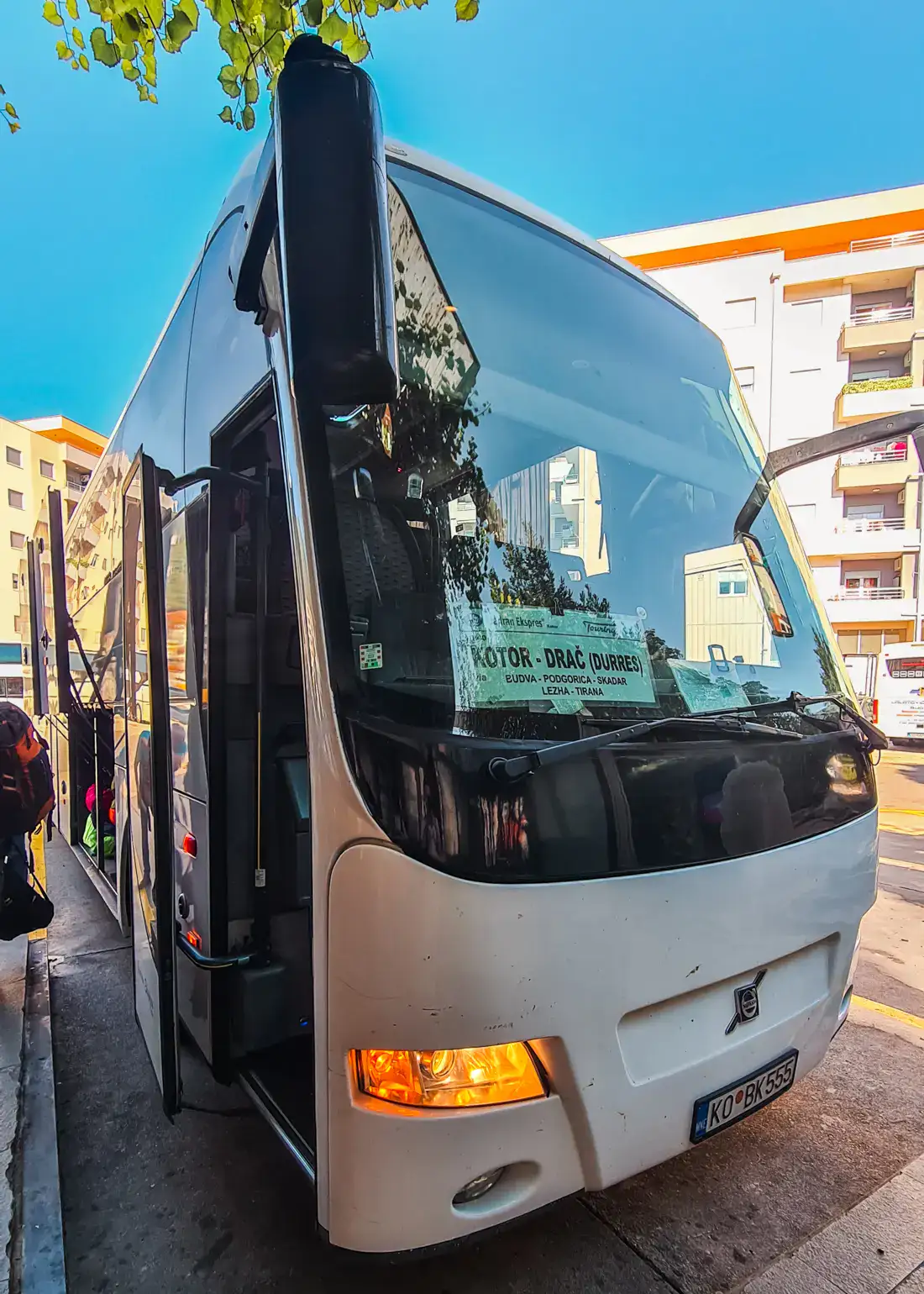 Close up of the front of a large white bus, with Budva - Durres sign in the window, on a sunny day with blue skis, light yellow apartment buildings in the background and Budva Bus station.