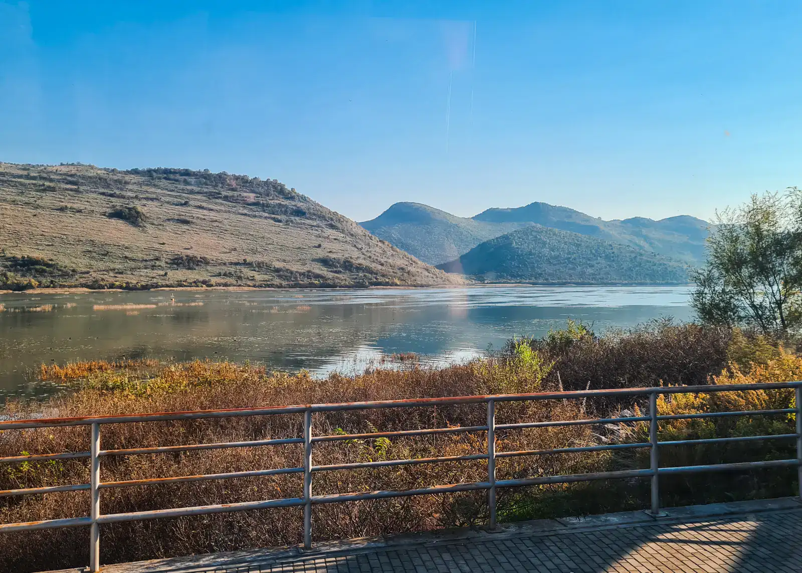 View over metal fence, of a calm lake surrounded by fall colored greenery, hills and mountains in the background, while waiting at the border between Montenegro and Albania.