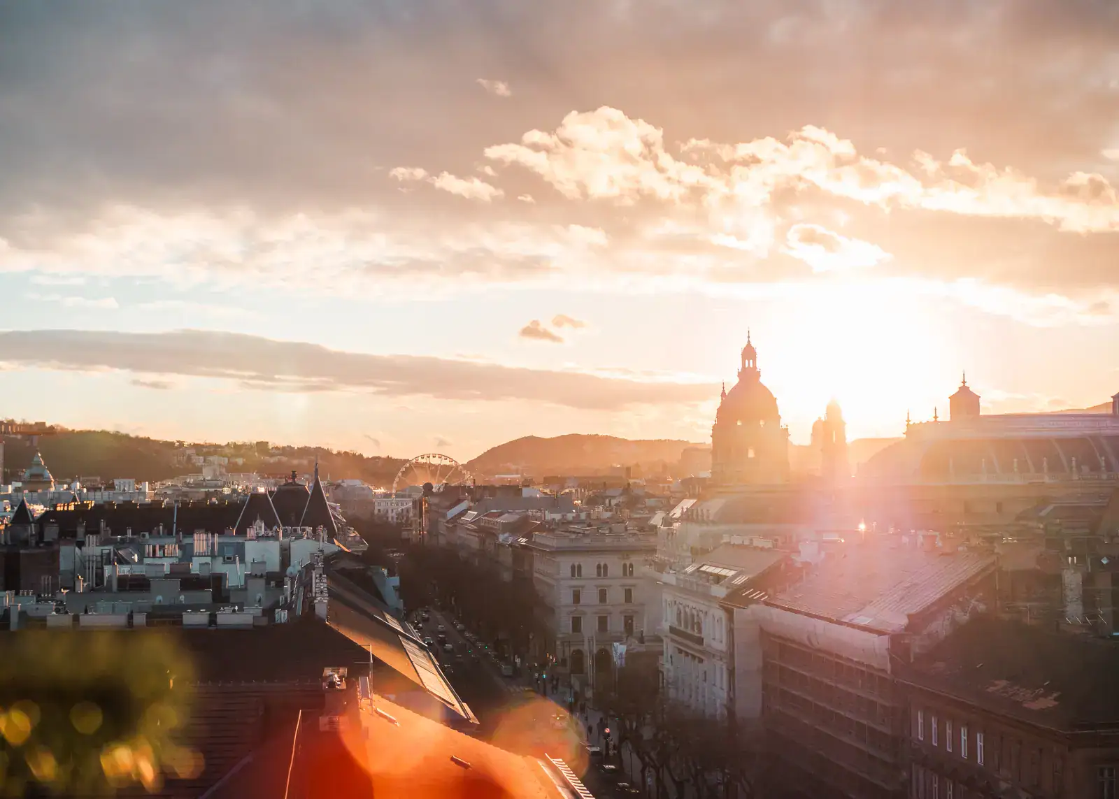 Sunset view from 360 rooftop bar in Budapest, orange light on a cloudy sky, looking over old buildings on a church dome and a Ferris wheel in the distance. A must during Budapest Itinerary.