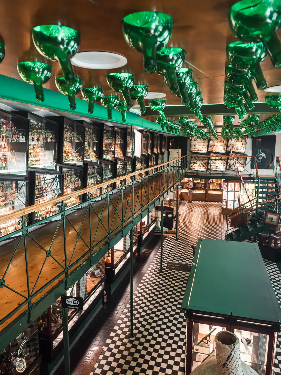 View from the second floor of the Unicum Museum, looking down on a black and white checkered floor, rows of green bottles on the ceiling, glass cases of old bottles along the wall, and alternative on your Budapest itinerary.