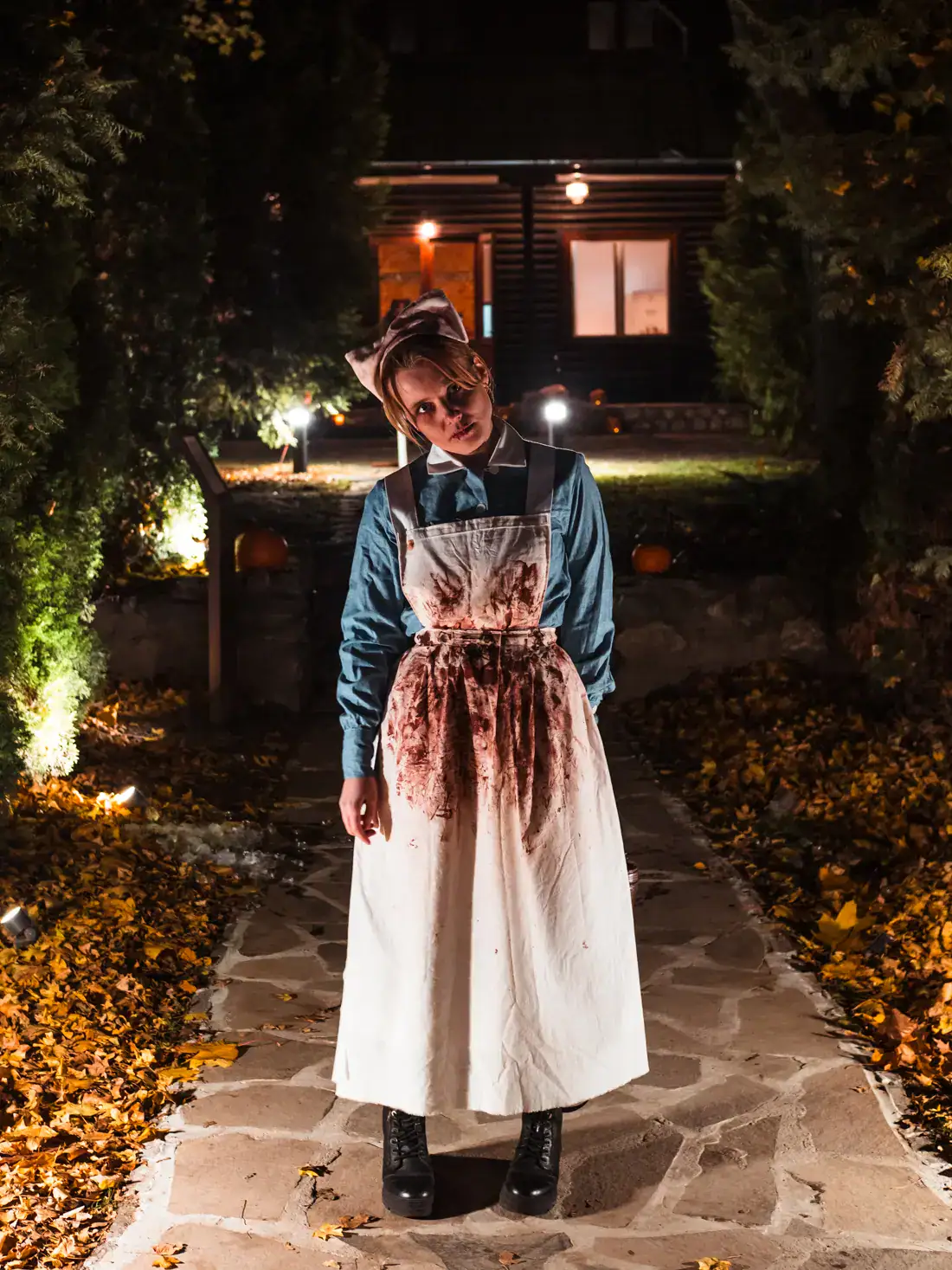 Woman wearing an old nurse uniform, grey dress with a white apron and hat, covered in blood, with hear head tilted, standing on a stone walkway with lights and leaves along the side at Bran Castle Halloween Party.
