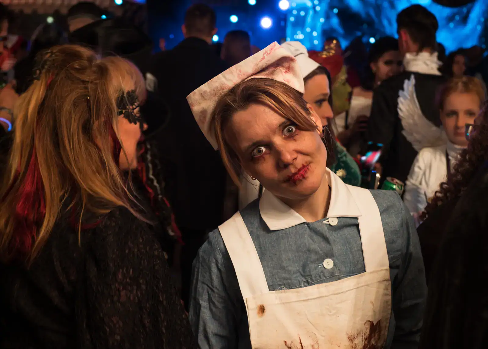 Woman wearing an old nurse costume, grey dress with white apron and hat, smeared in blood, looking with crazy, surrounded by people in costumes and blue light in the background, Halloween party at Bran Castle in Transylvania.