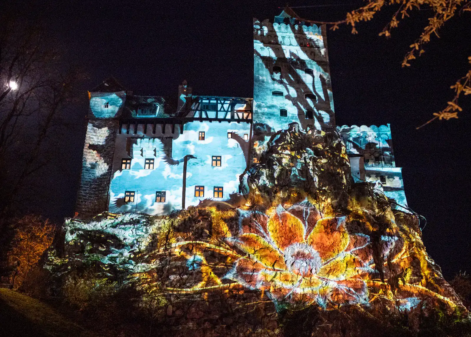 Looking up at the old, stone, Bran Castle lit up by a blue light show with a large yellow lotus flower, with a black sky in the background, during the Halloween celebrations.