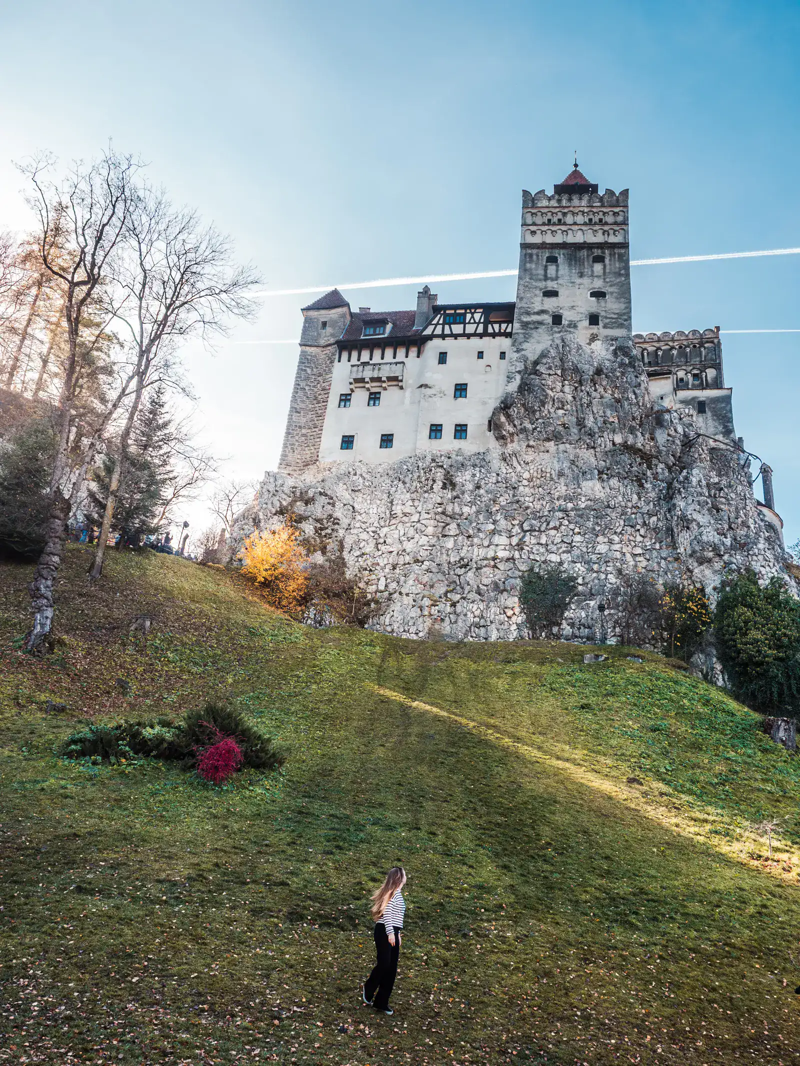 Woman wearing a white sweater and black pants, walking up a green hill towards Bran Castle perched on a rock on a sunny day in Romania.