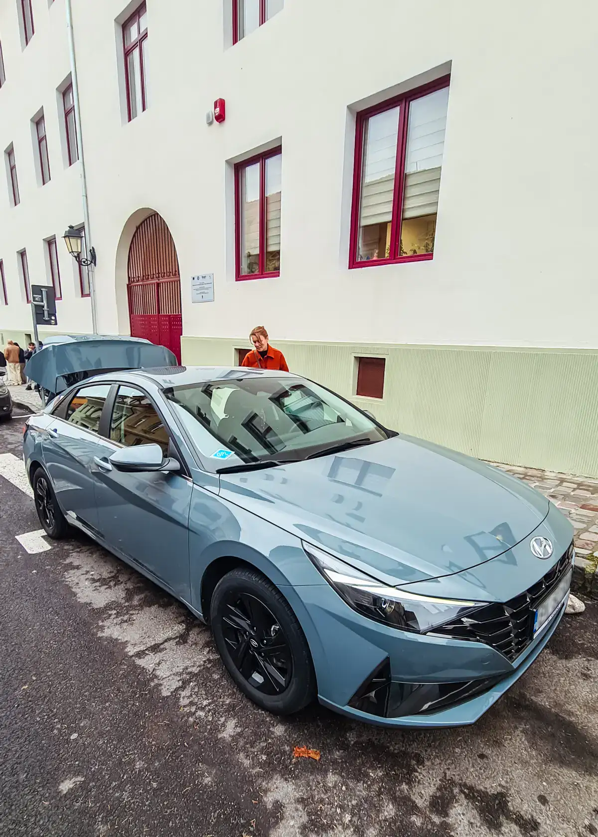 Woman wearing an orange jacket standing behind a light blue Hyundai, next to a light green apartment building, before a private transfer with Daytrip from Brasov to Bucharest.