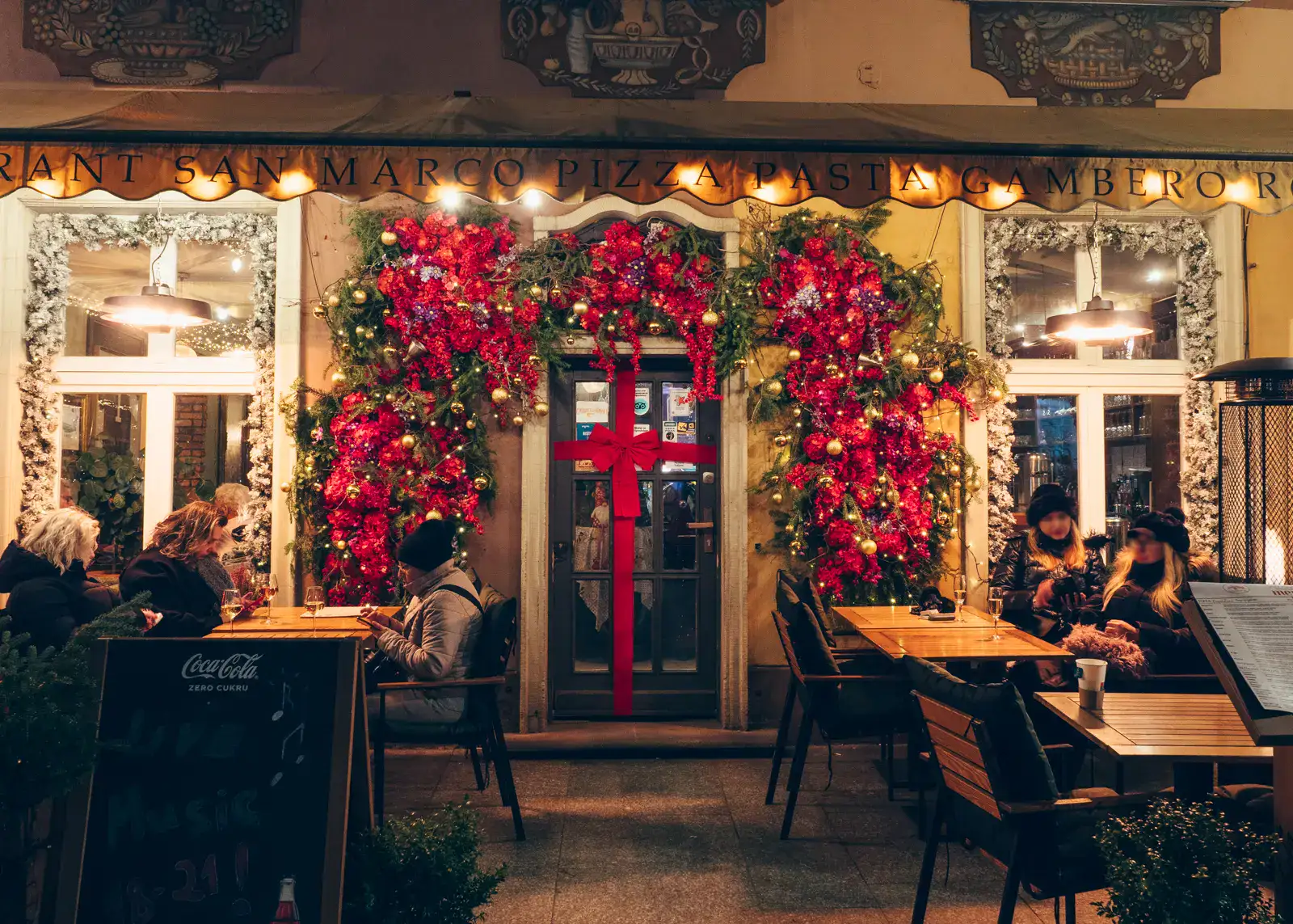 Grey restaurant door decorated with a red ribbon, red flowers, gold baubles and greenery around it, a few people in coats sitting at the tables outside in Gdansk for Christmas.