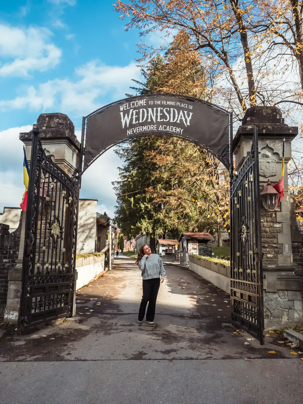 Woman wearing a grey sweater and black pants, flicking her hair, under a metal gate with a sign reading, filming location for Wednesday Nevermore Academy, on a private transfer from Brasov to Bucharest with Daytrip.
