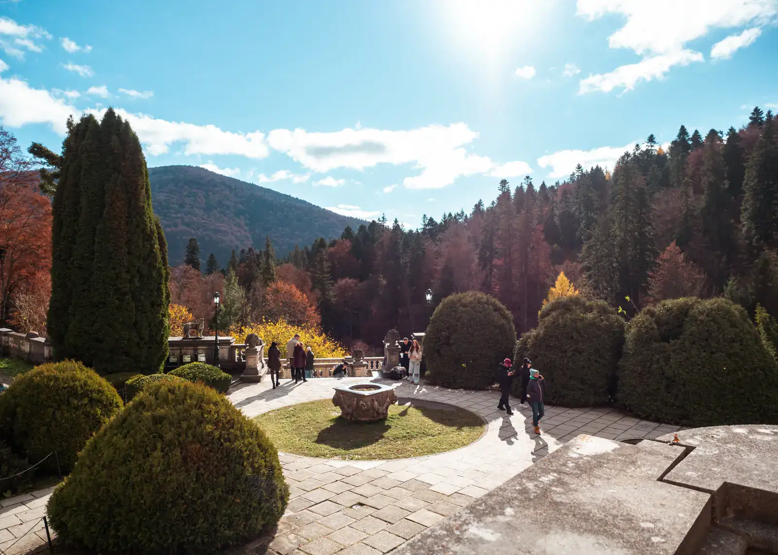 View from Peles Castle of the gardens with a stone fountain, surrounded by rounded bushes, mountains in the background with orange, red and green trees on a sunny day, transfer with Daytrip.