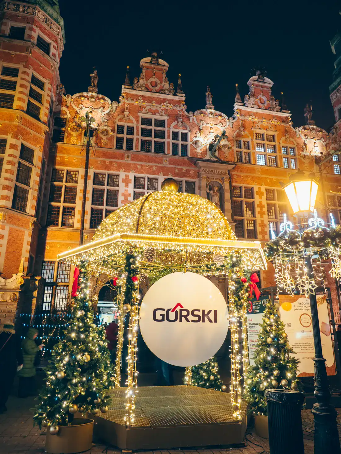 Gazebo covered in lights with a small Christmas tree on both sides, black street light on the right, old, ornate pinkish building in the background at Gdansk Christmas Market.
